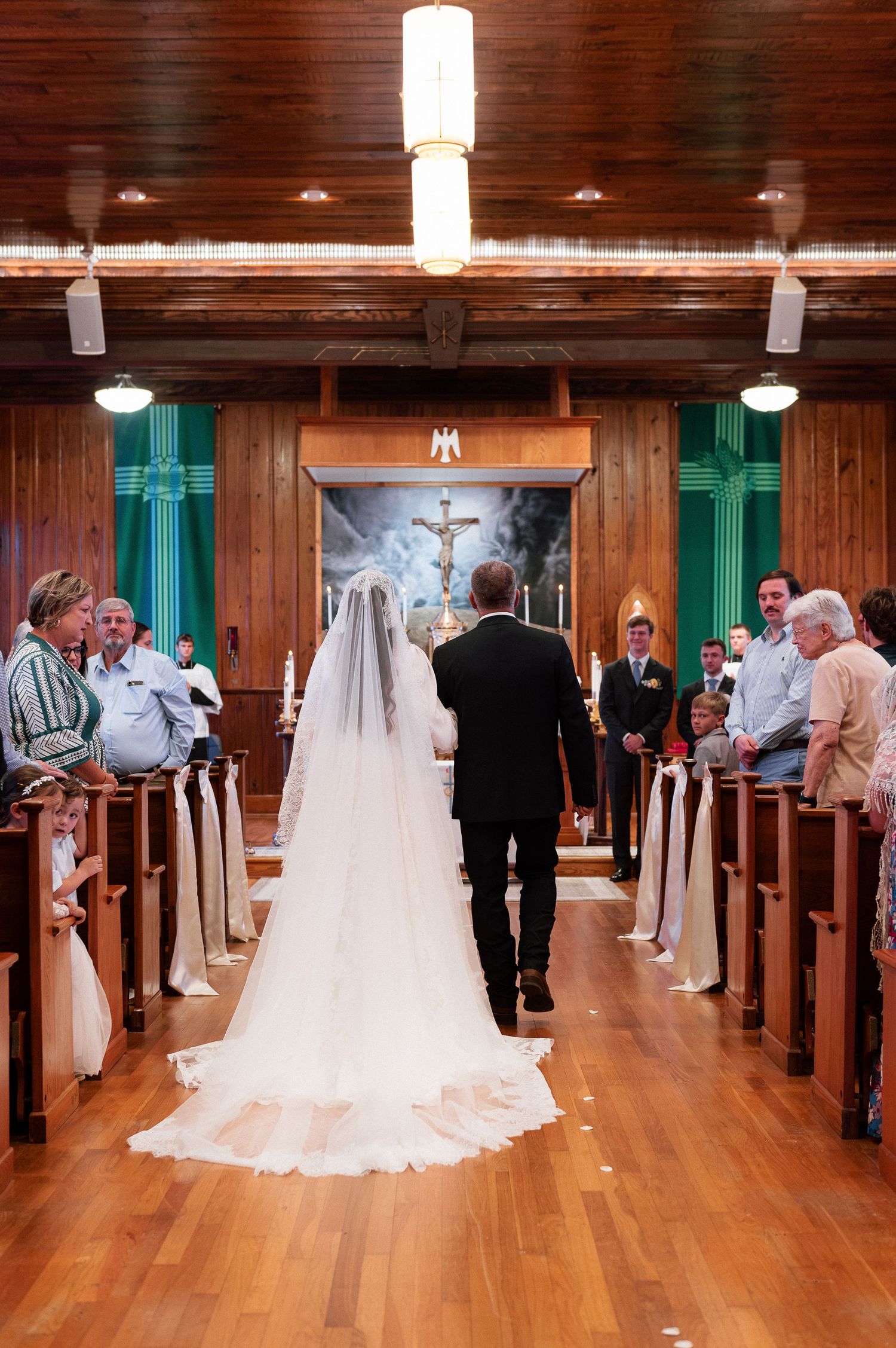 Bride walking down the aisle with escort in a wooden church with green cross accents during wedding ceremony.