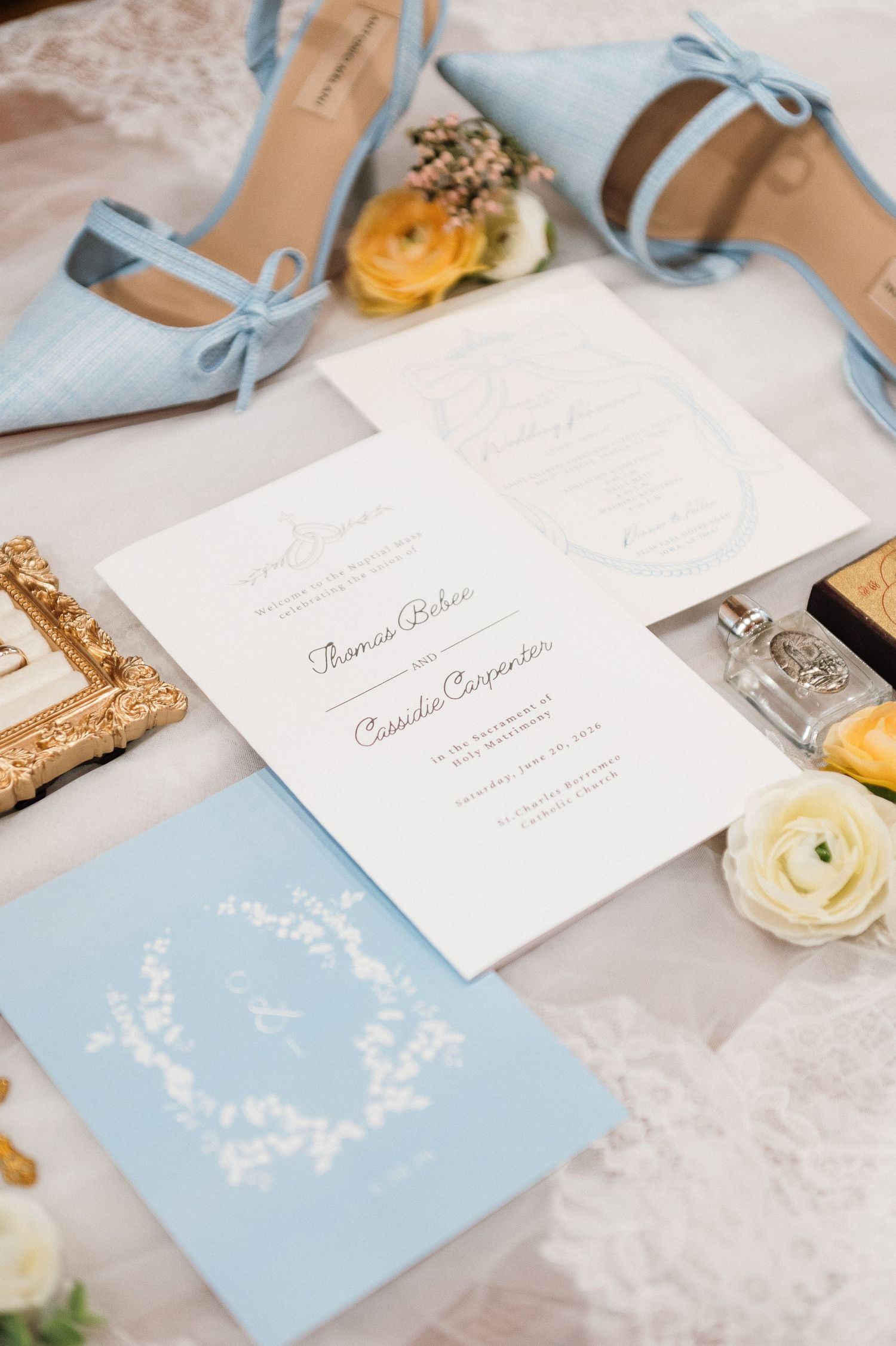 Elegant wedding invitation suite with light blue heels, white stationery, floral accents, and gold details on a soft backdrop.