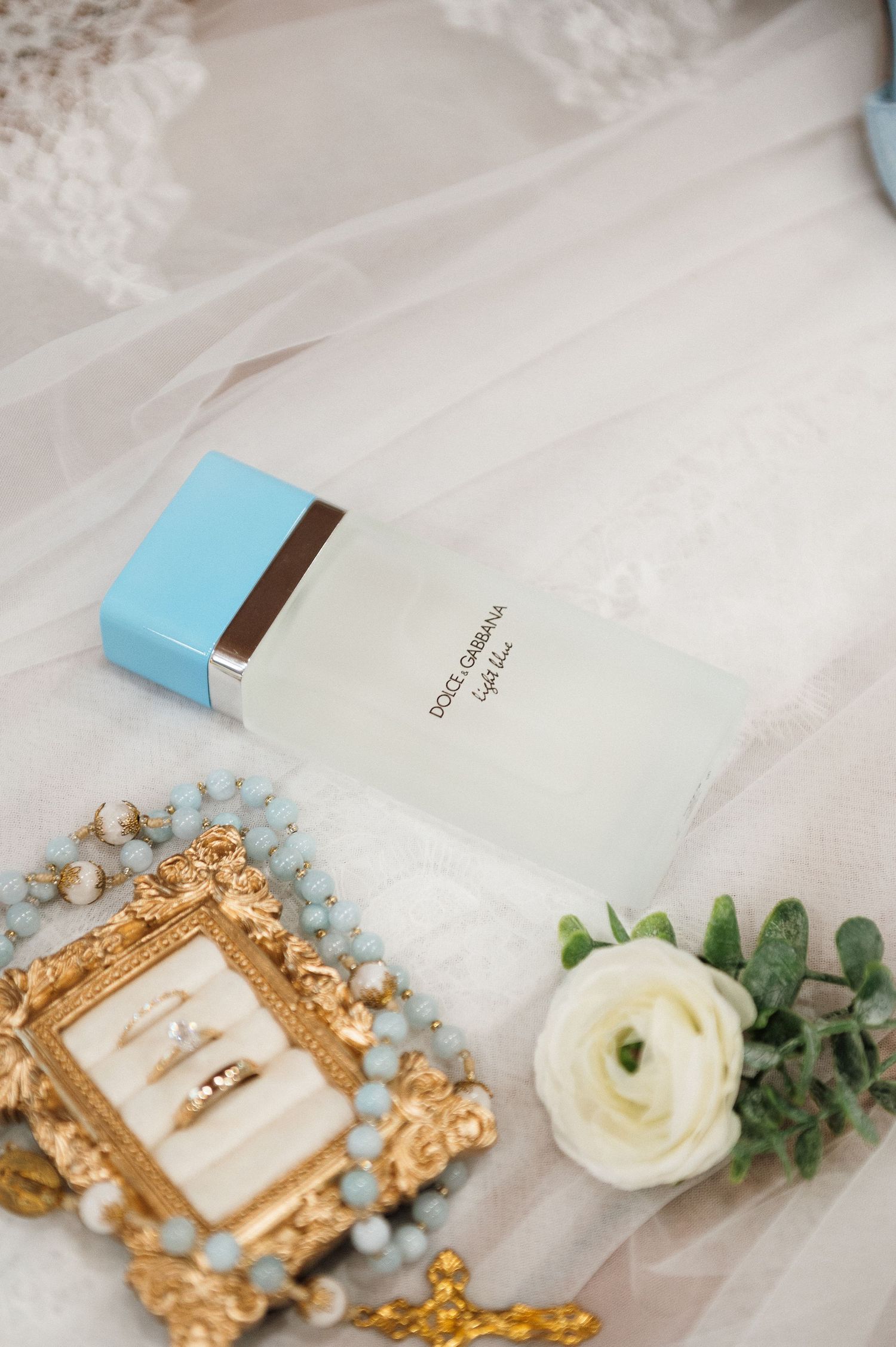 Dolce & Gabbana Light Blue perfume beside gold ring box, pearl necklace, and white ranunculus flower on bridal veil.