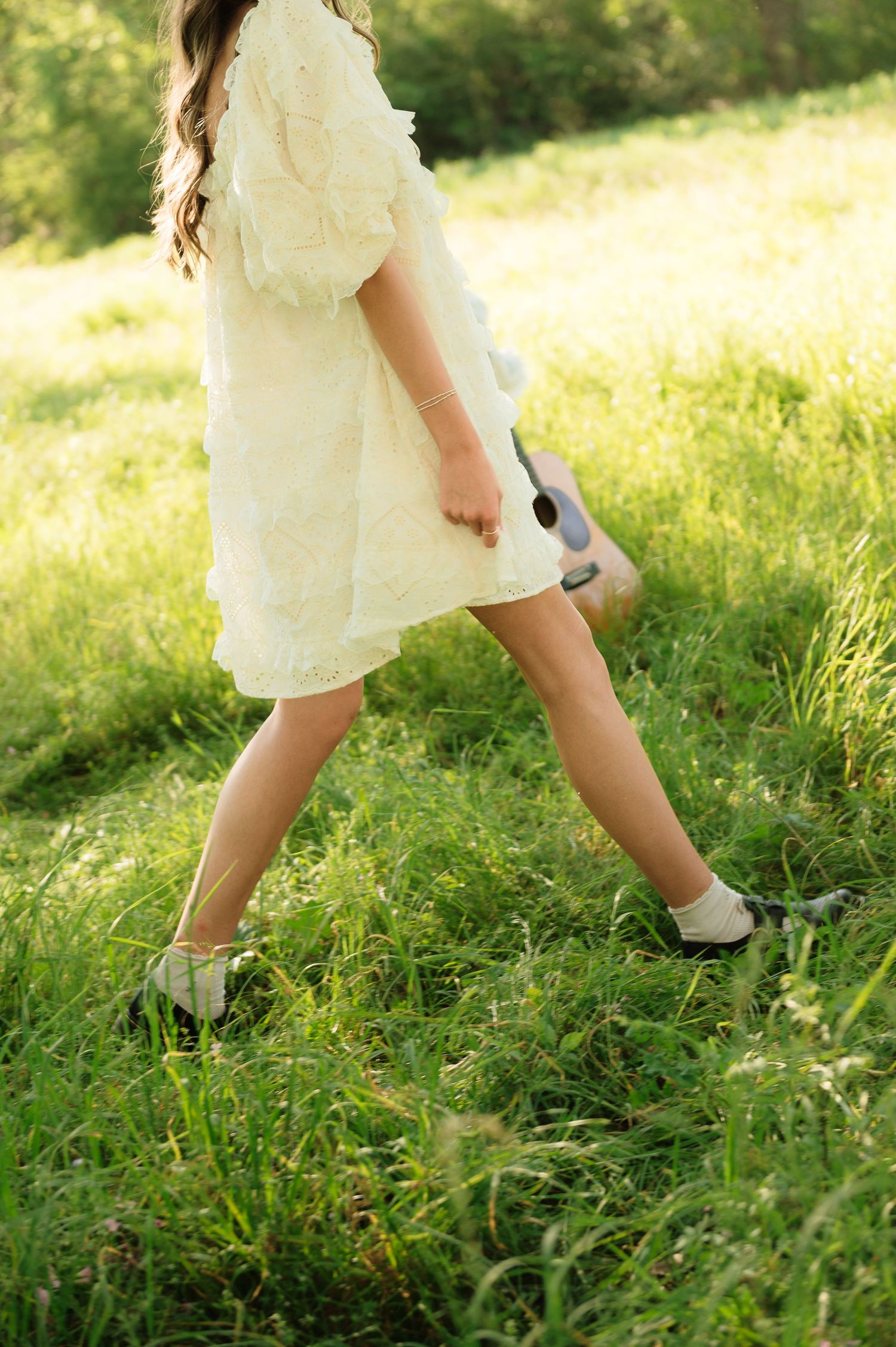 A young girl in a white lace dress walks through a sunny green meadow, carrying a small guitar.