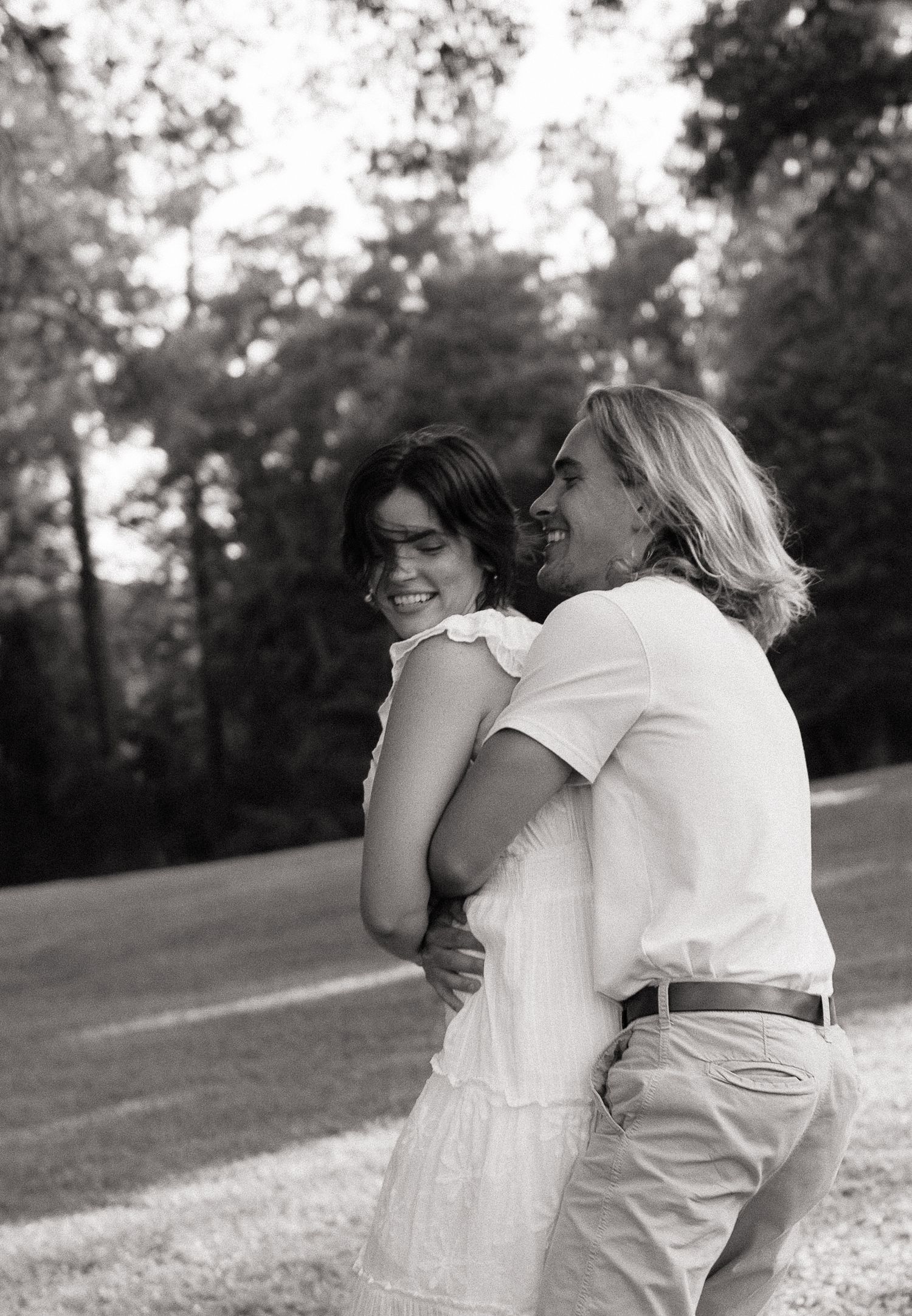 Black and white photo of a laughing couple embracing outdoors in a grassy park with trees in the background.