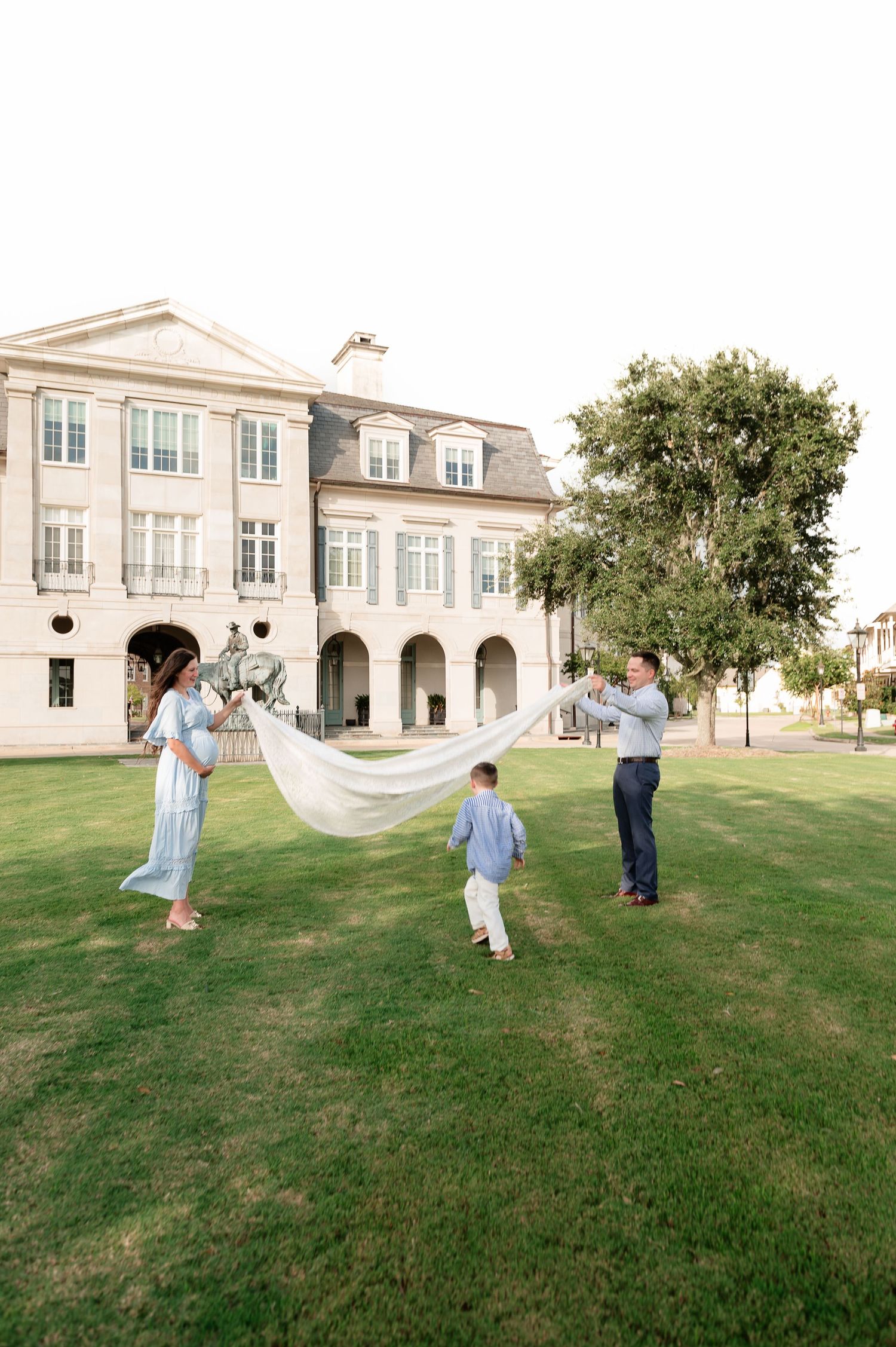 Family of three holds a flowing white fabric on a green lawn in front of a grand cream-colored mansion during an outdoor photo session.