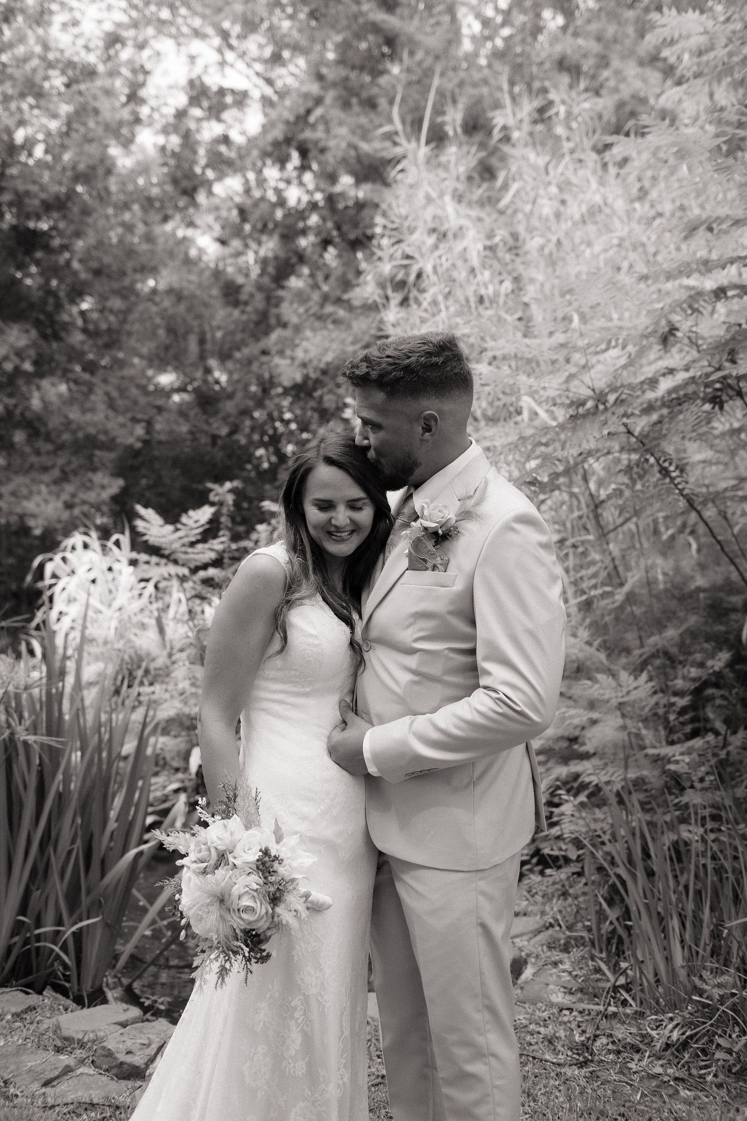 Black and white photo of a bride and groom embracing in a lush garden, bride holding a bouquet.