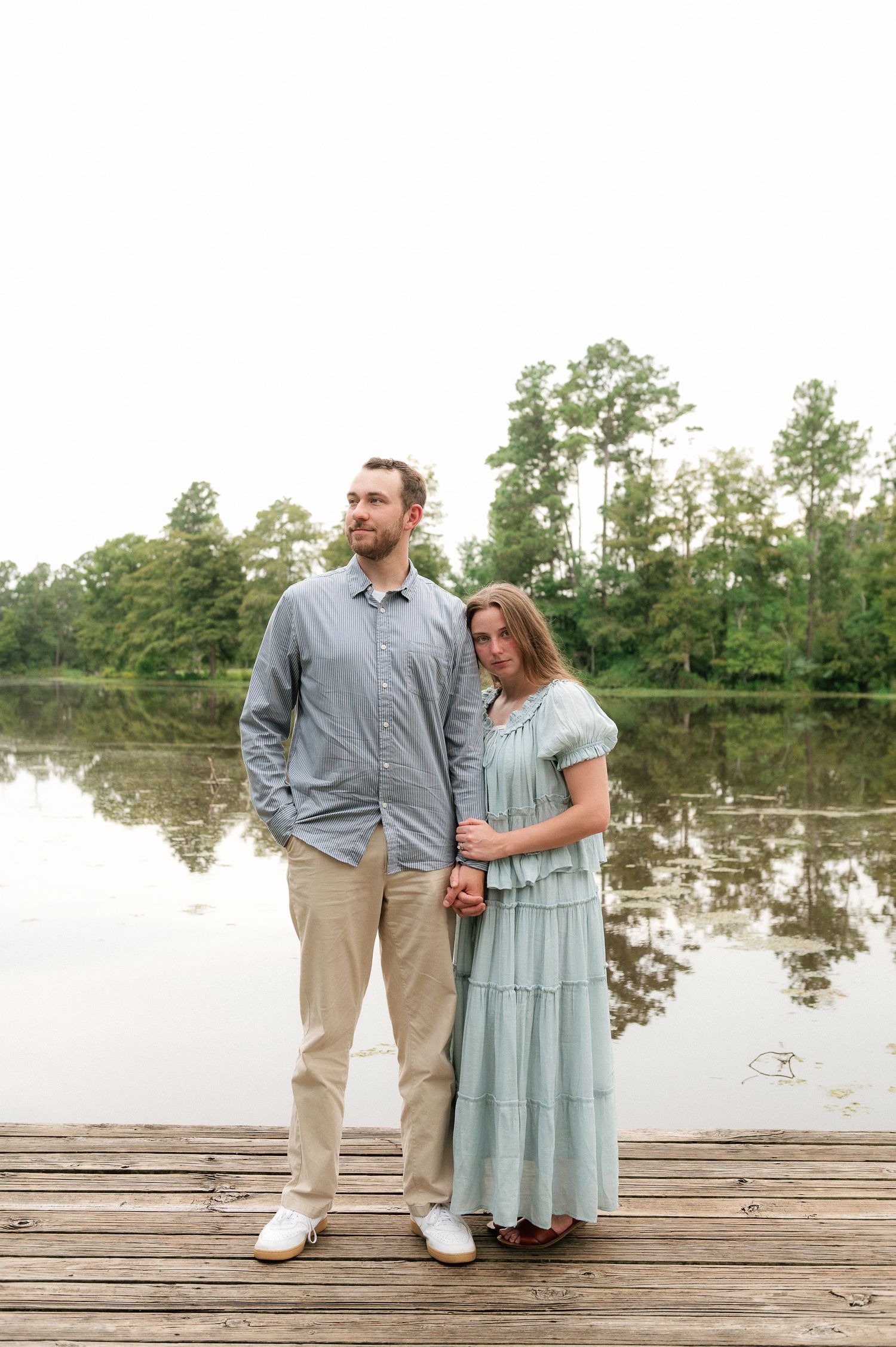 Couple standing on a wooden dock by a calm lake, man in blue shirt and woman in mint dress, surrounded by trees.