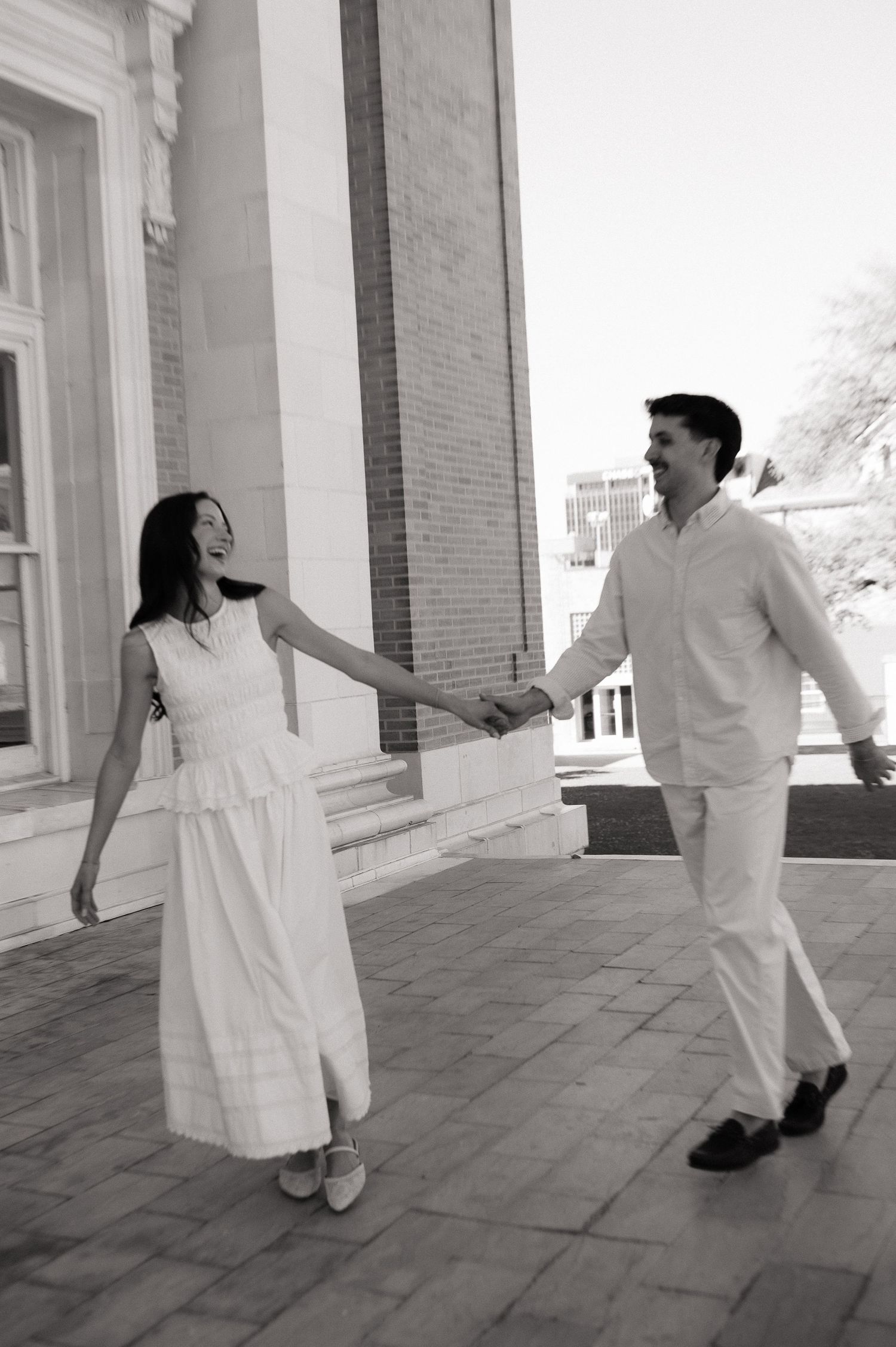 Black and white photo of a joyful couple holding hands and walking together outside a classic building.