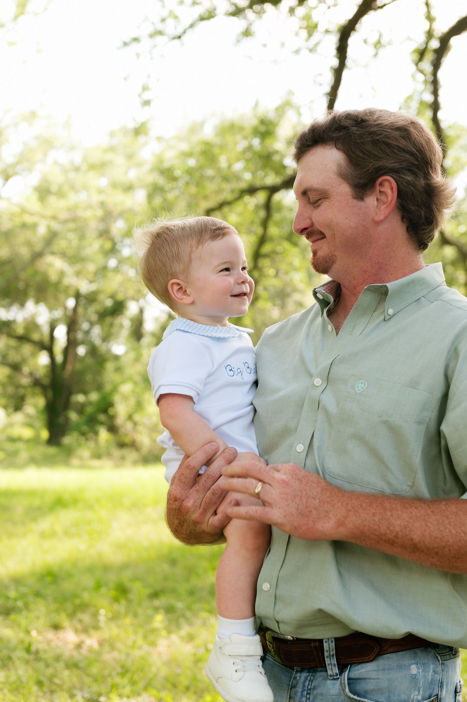 A smiling man in a green shirt holds a happy baby boy outdoors in a lush green park setting.