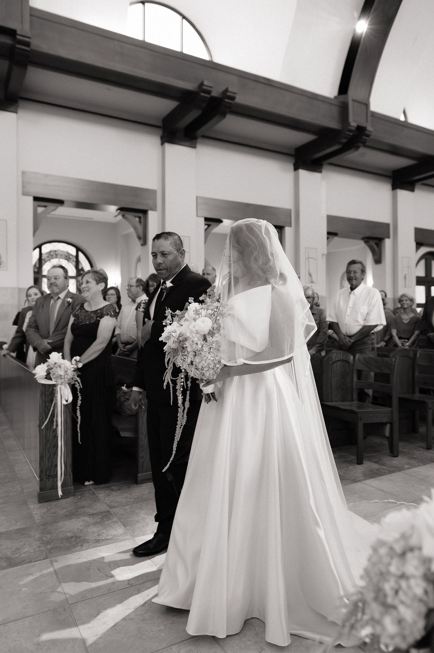 Bride in white gown and veil walks down church aisle with escort holding bouquet at wedding ceremony.