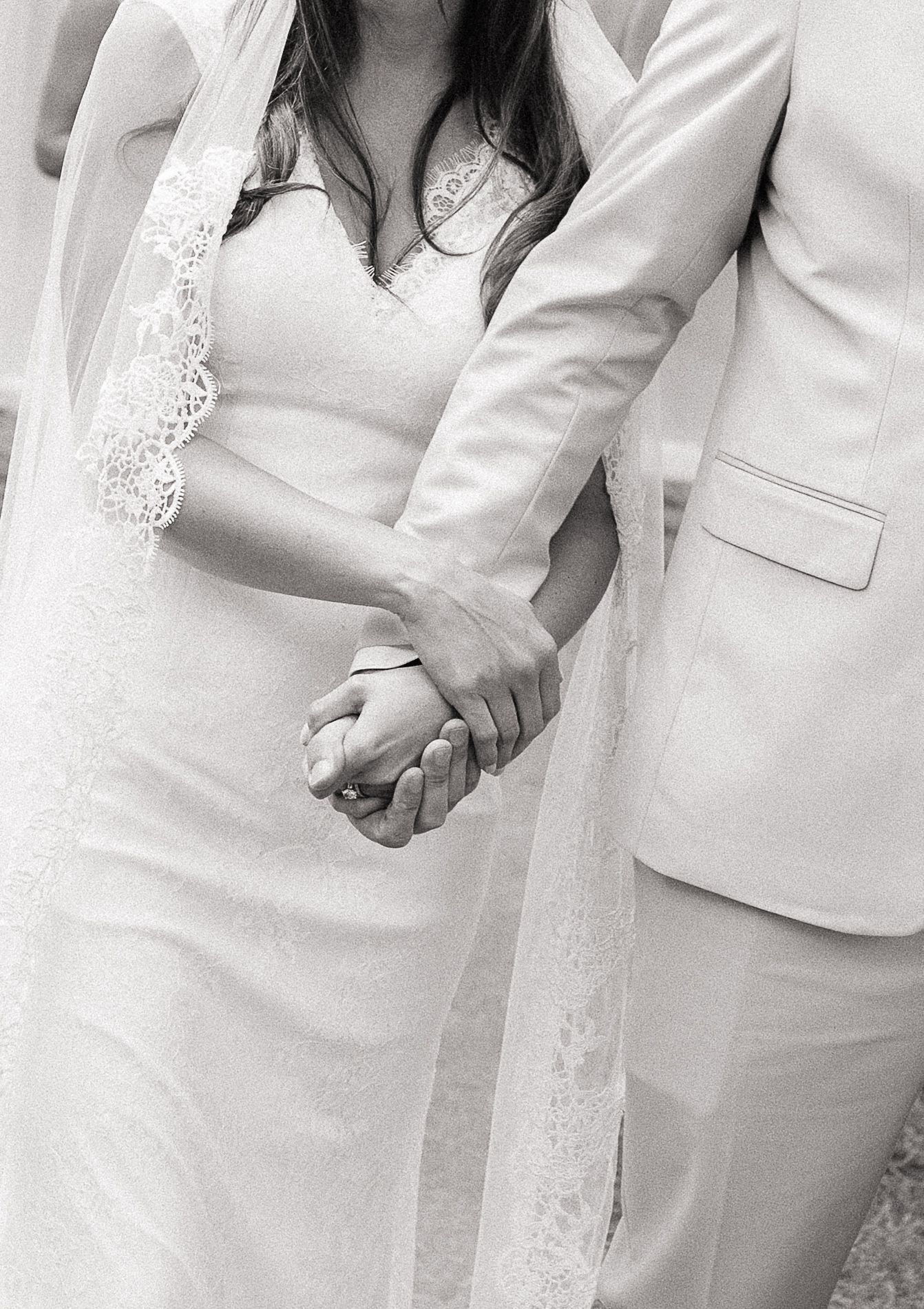 Black and white photo of bride and groom holding hands, showcasing wedding dress, lace veil, and light suit.