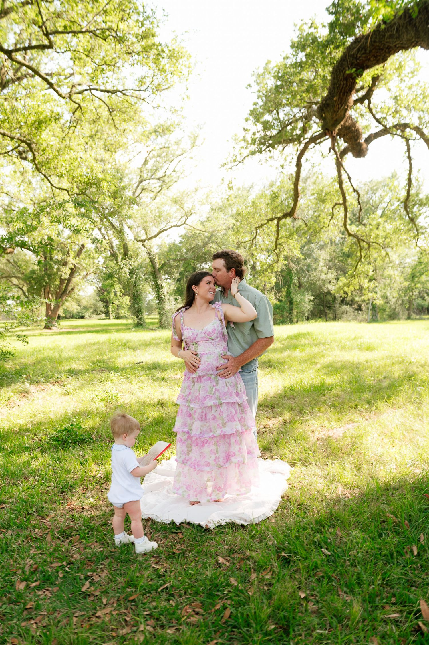 Pregnant woman in pink floral dress with husband and toddler during maternity photoshoot in park.