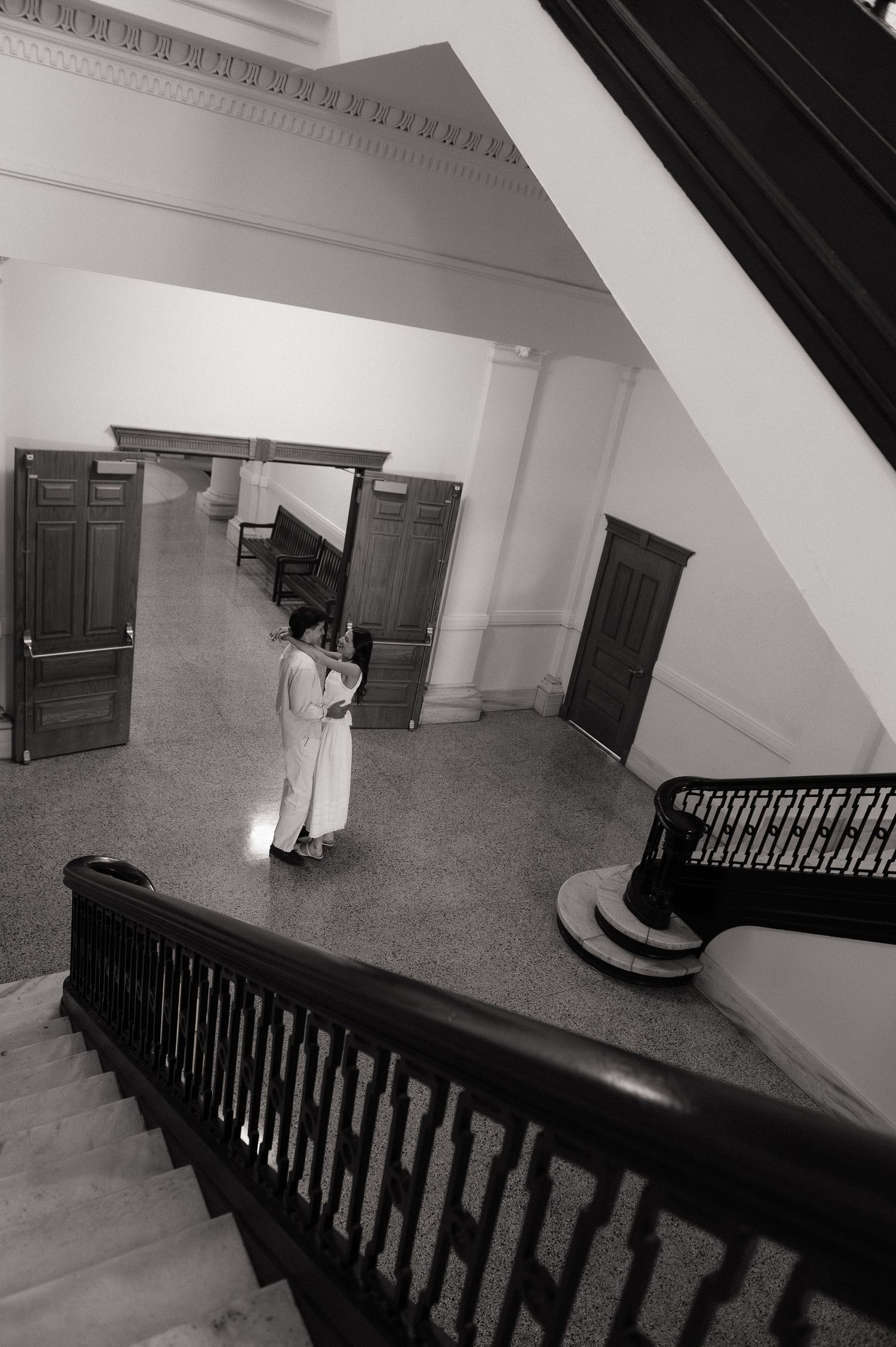 Black and white photo of a person standing in a grand building foyer, viewed from above a staircase with ornate railings.