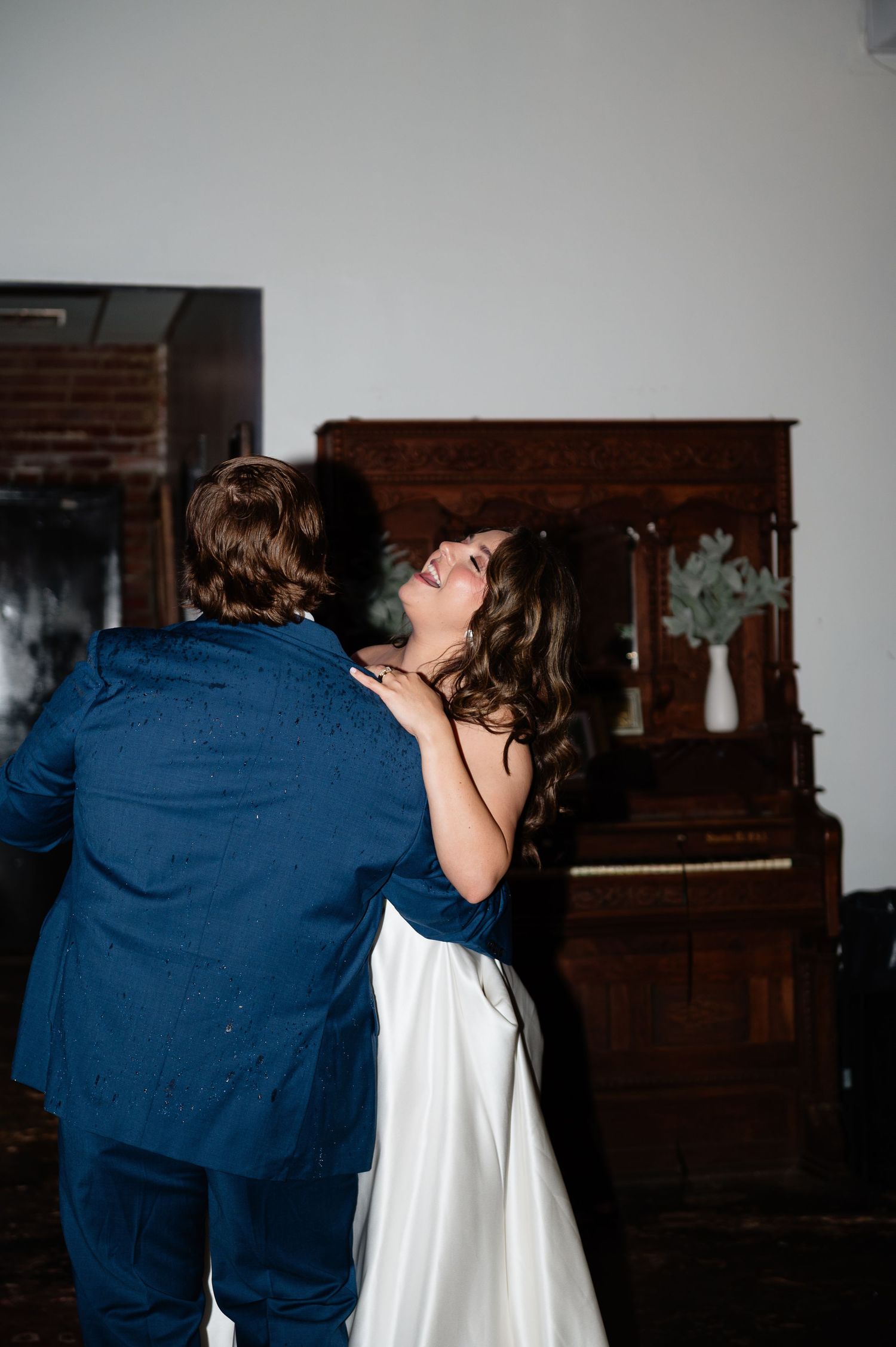 A bride in a white gown and groom in a blue suit share a romantic dance near a vintage upright piano.