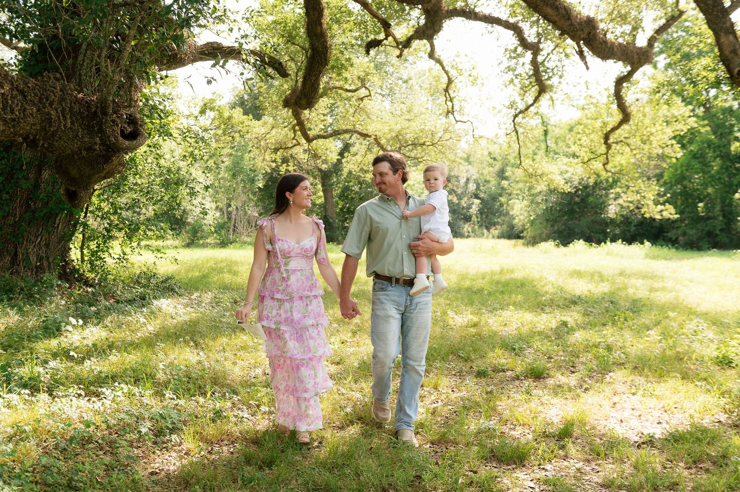 Family of three walking through lush green park, mother in floral dress, father holding baby, under oak trees.