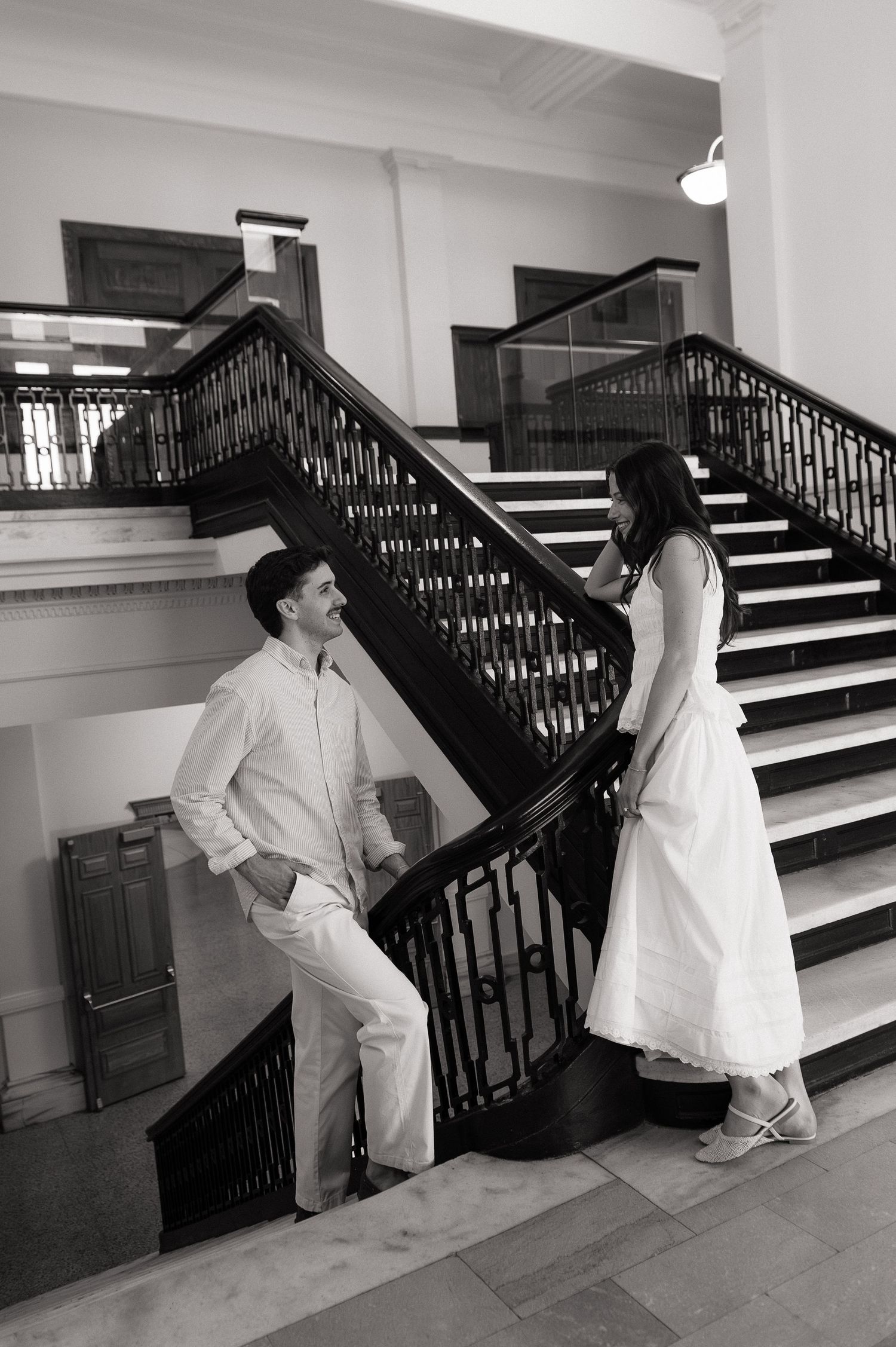 Black and white photo of a couple in white attire sharing a romantic moment on an elegant staircase.