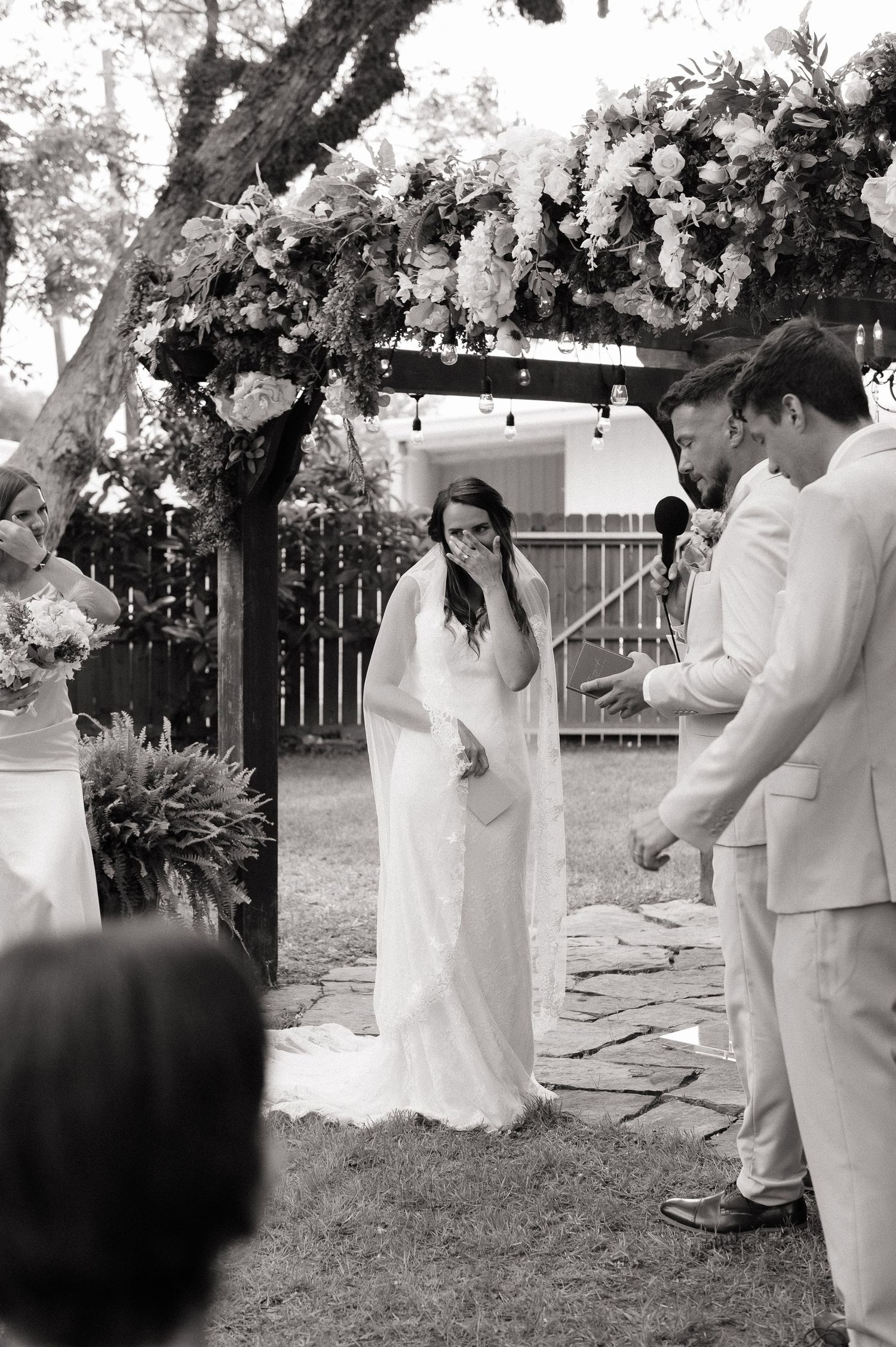 Bride laughing emotionally at outdoor wedding ceremony under floral arch with groom in tan suit.