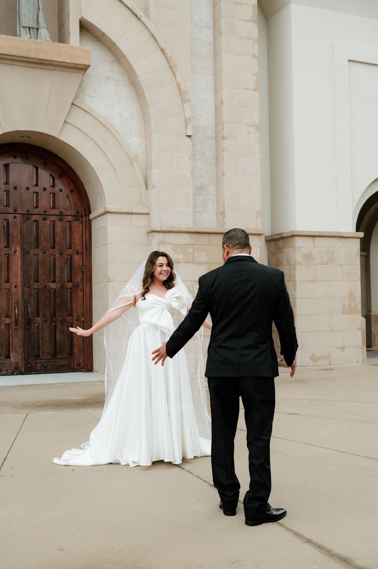 Bride in white gown with bow reacts joyfully during first look with groom in black suit outside stone church.