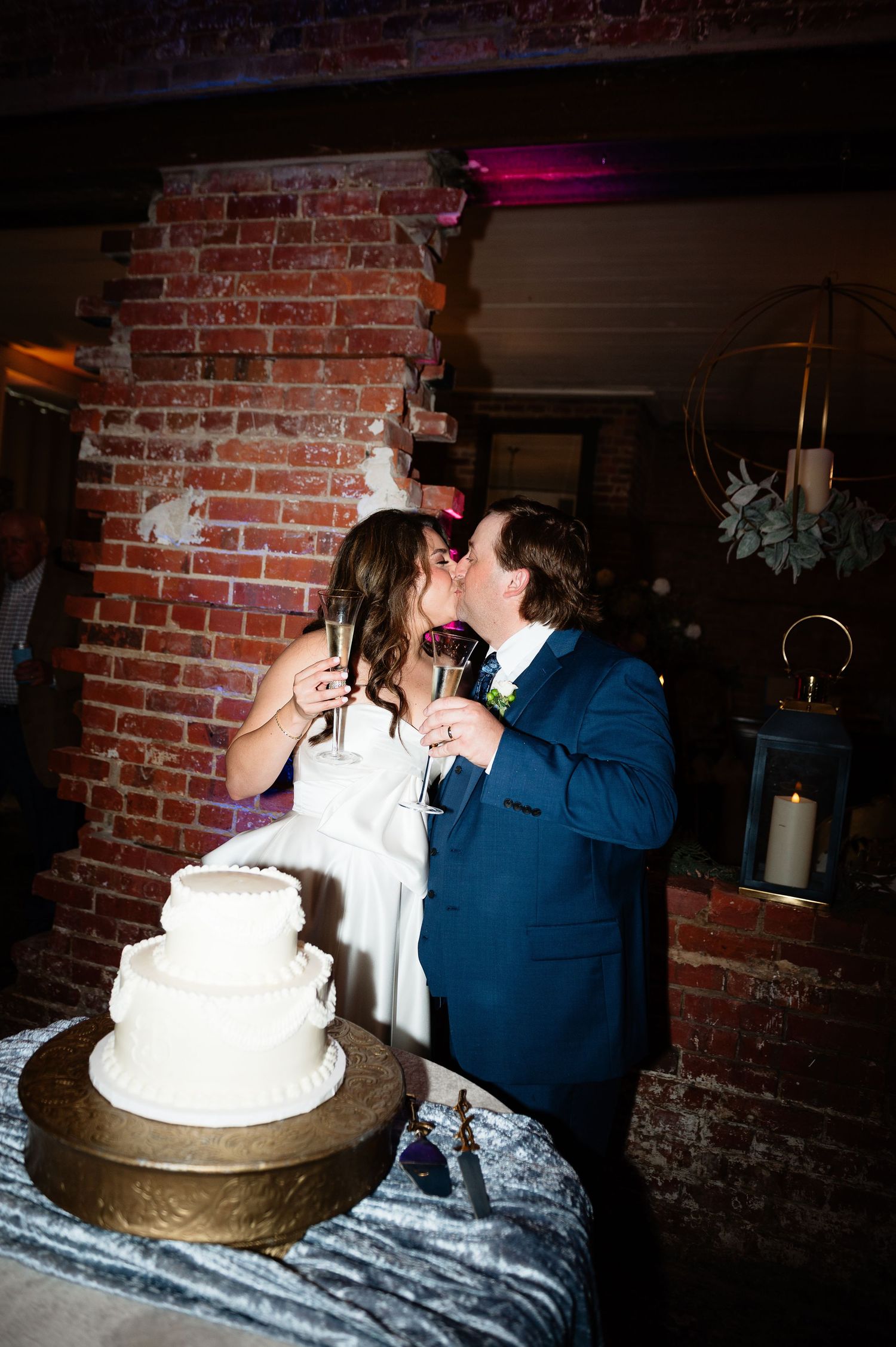 Newlyweds share romantic kiss over white three-tier wedding cake at rustic brick wall reception venue.