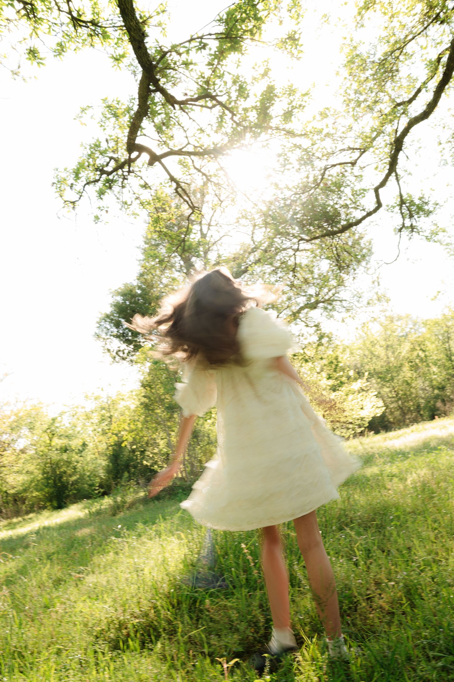 A young girl in a white dress twirls joyfully in a sunlit green meadow beneath a tree.