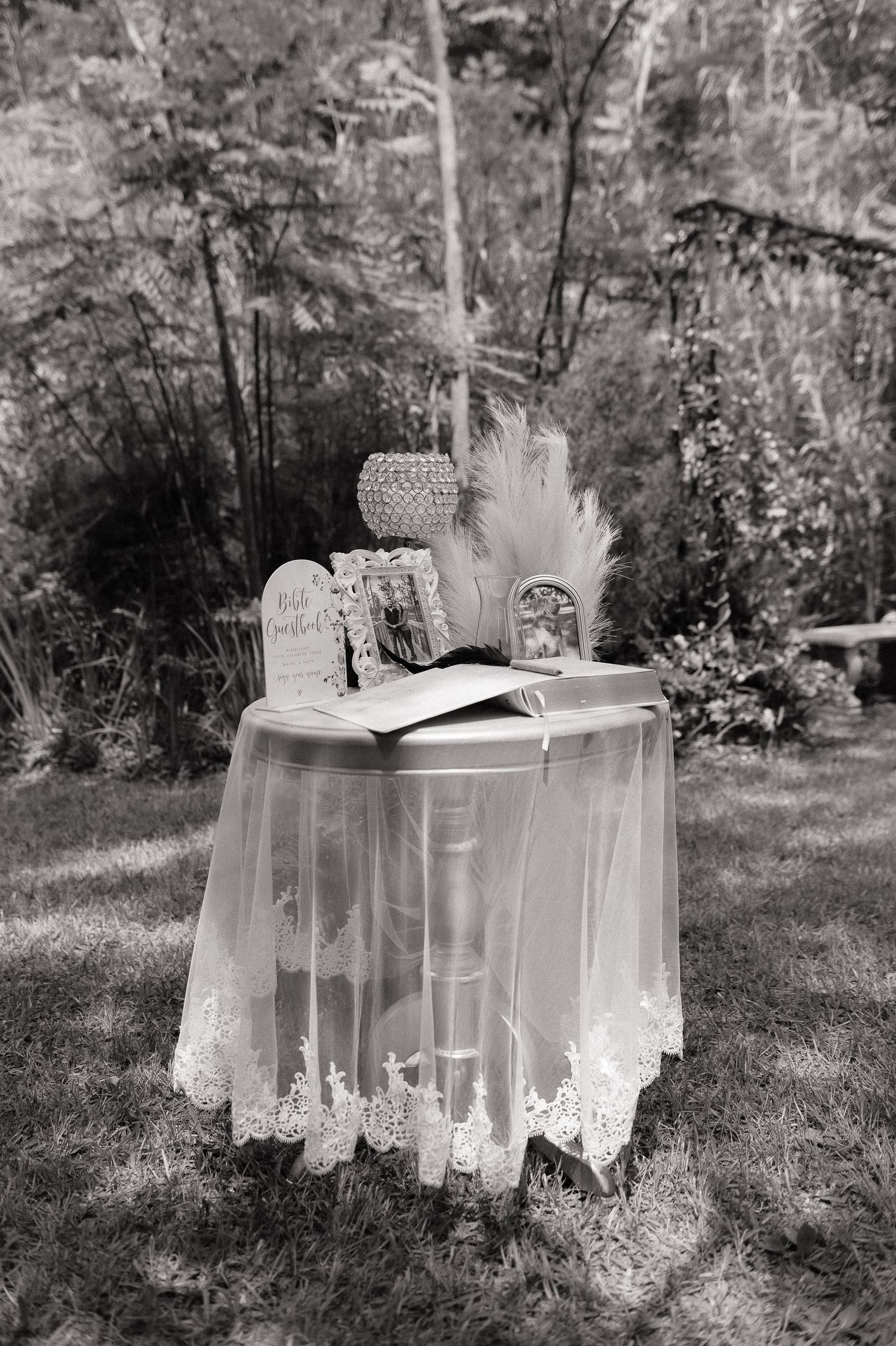 Black and white photo of an elegant lace-draped outdoor wedding table with an open guest book and floral decor.