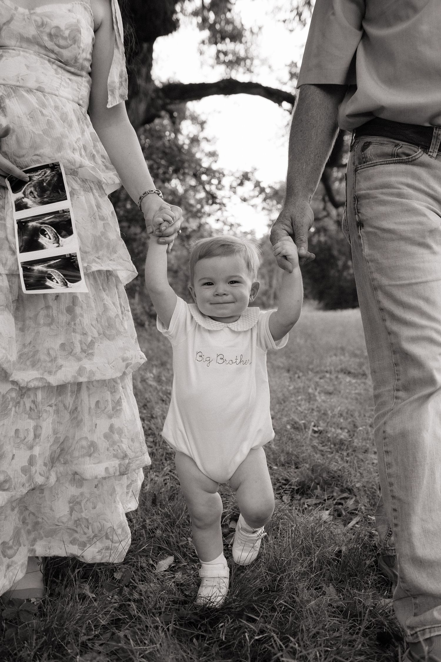 Black and white photo of toddler in Big Brother onesie held by parents hands outdoors, with ultrasound photos visible.