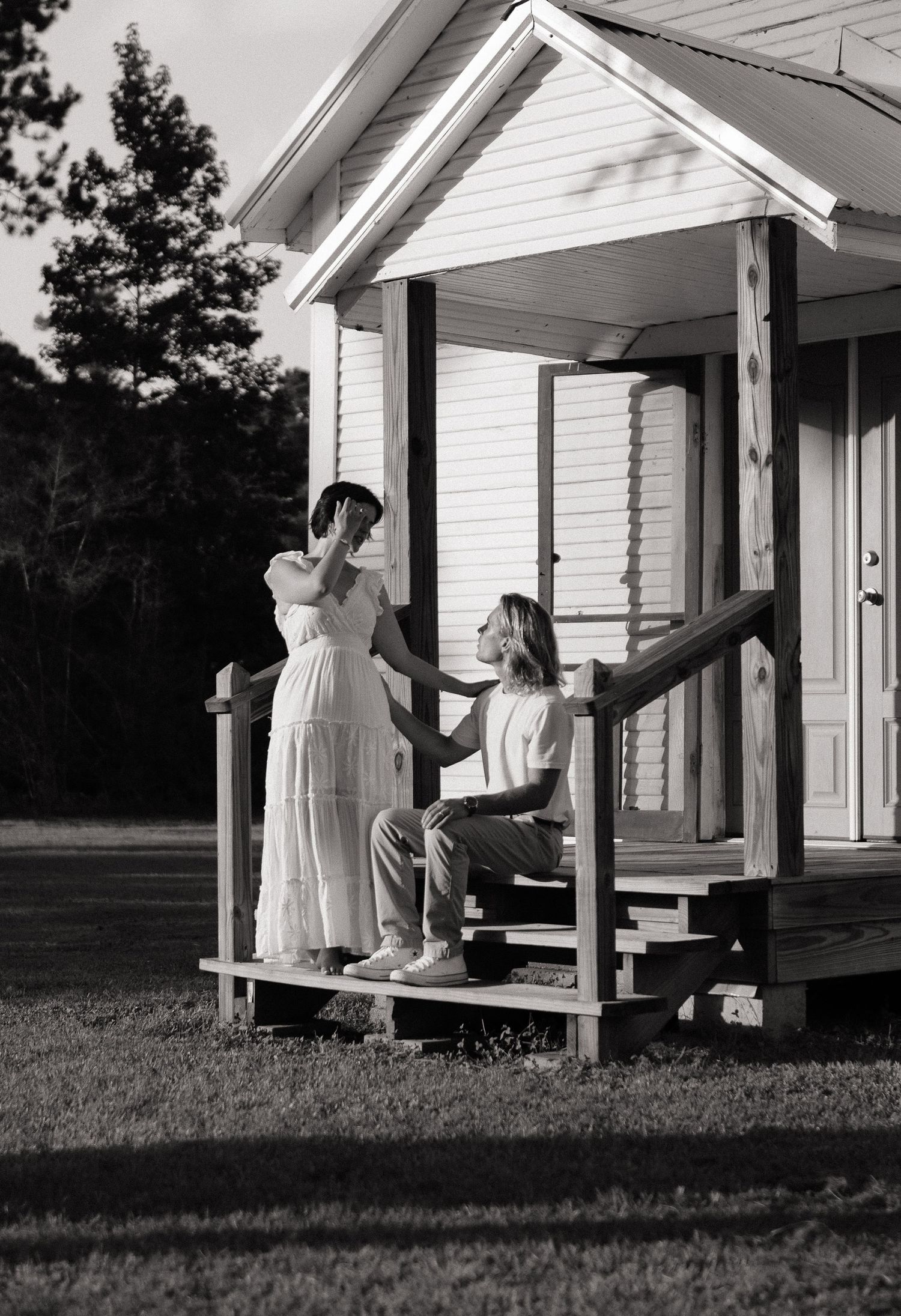 Black and white photo of a couple on a white cottage porch, woman in flowing dress, man seated on steps.