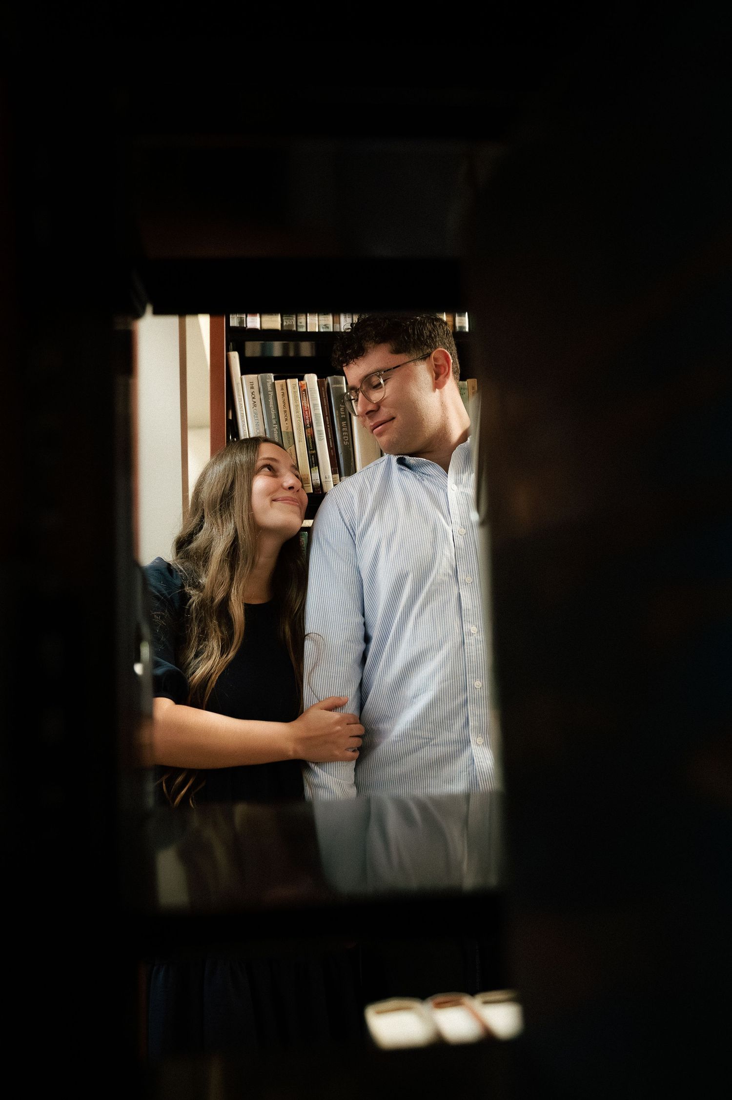 A couple shares a romantic moment in a cozy library, smiling at each other surrounded by bookshelves.
