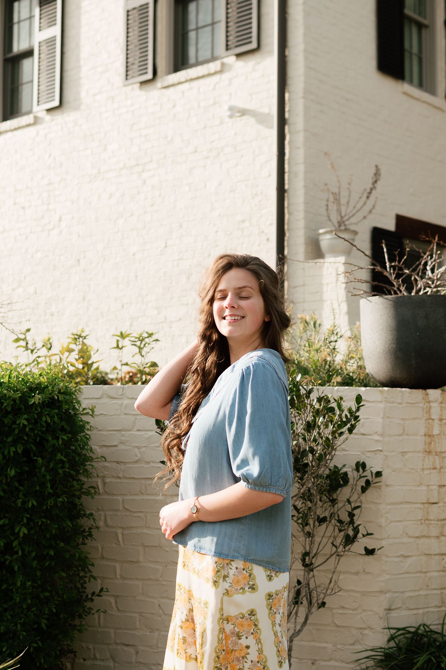 Young woman with long wavy hair smiling, wearing blue top and floral skirt, standing outdoors by white brick wall.