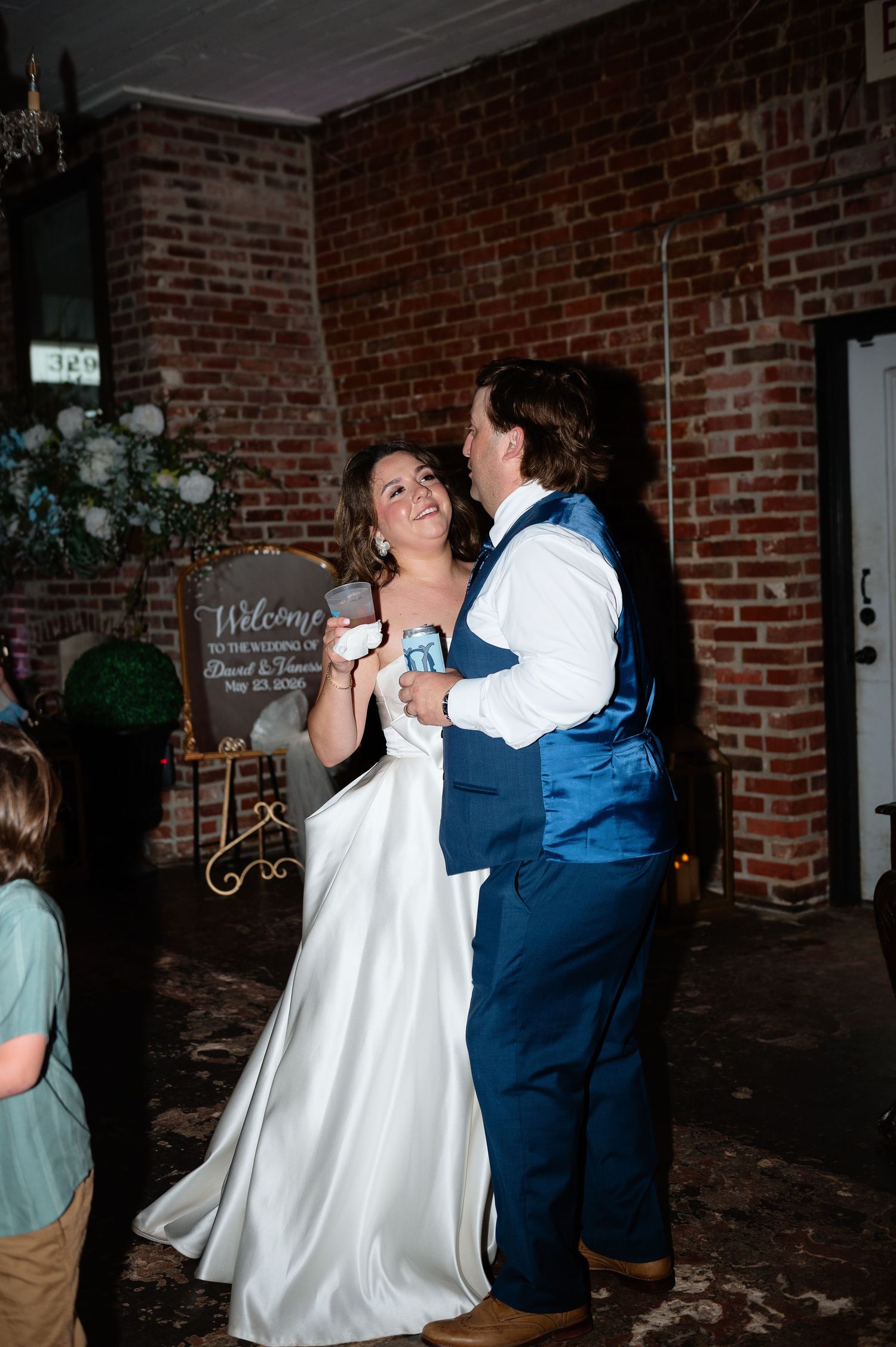 Bride in white gown and groom in blue vest share their first dance at a rustic brick-walled wedding venue.
