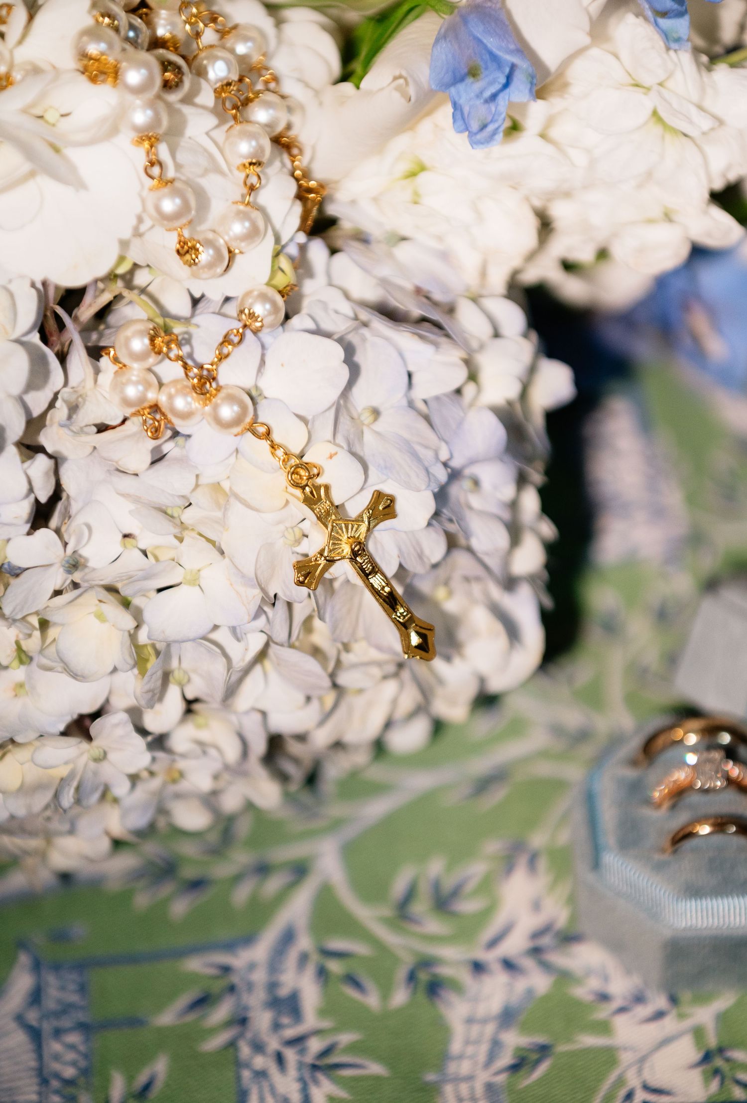 Gold cross rosary with pearl beads resting on white and blue hydrangea wedding bouquet with rings nearby.