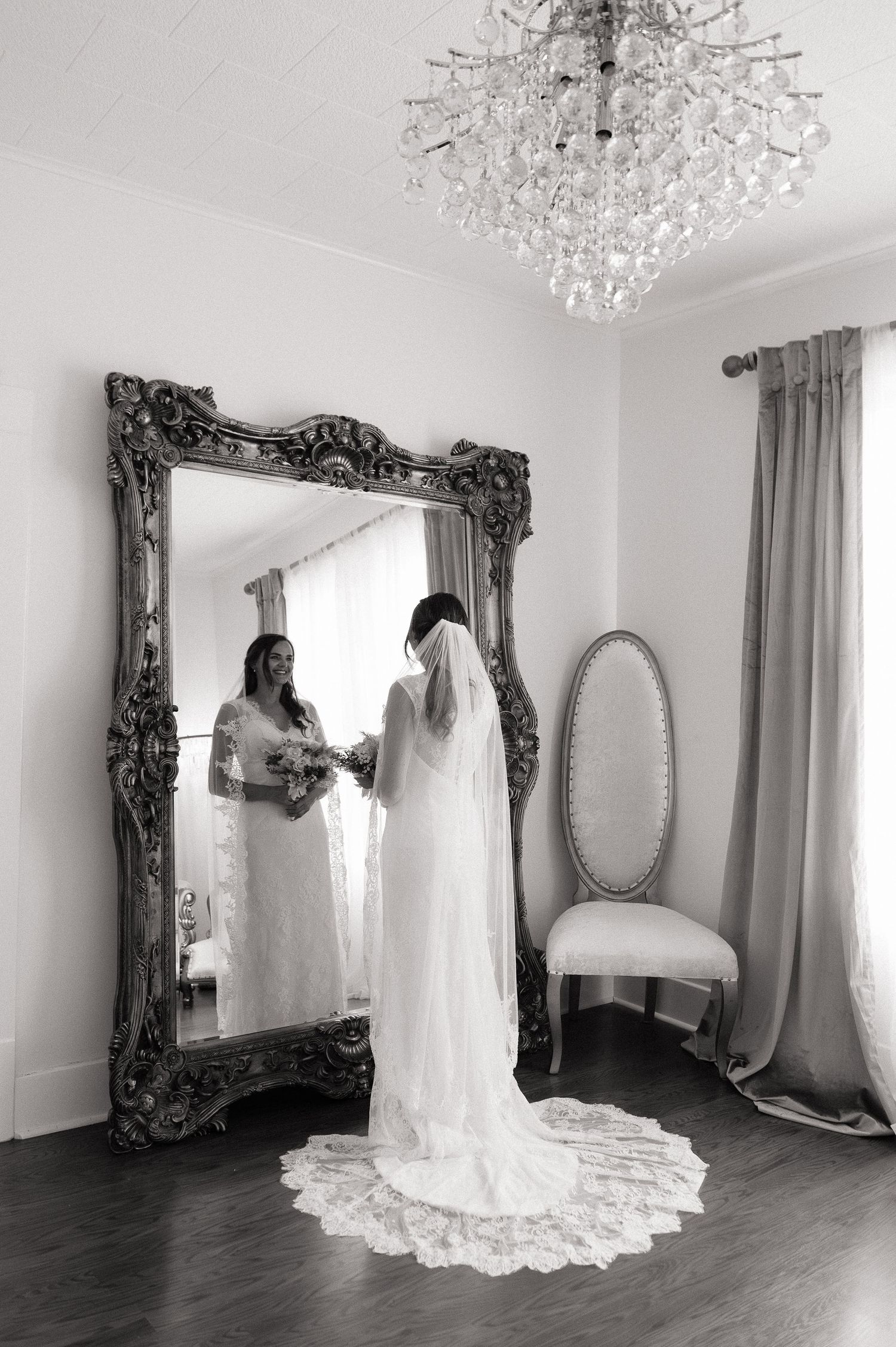 Bride in lace wedding gown with veil admiring her reflection in ornate silver mirror, crystal chandelier above.