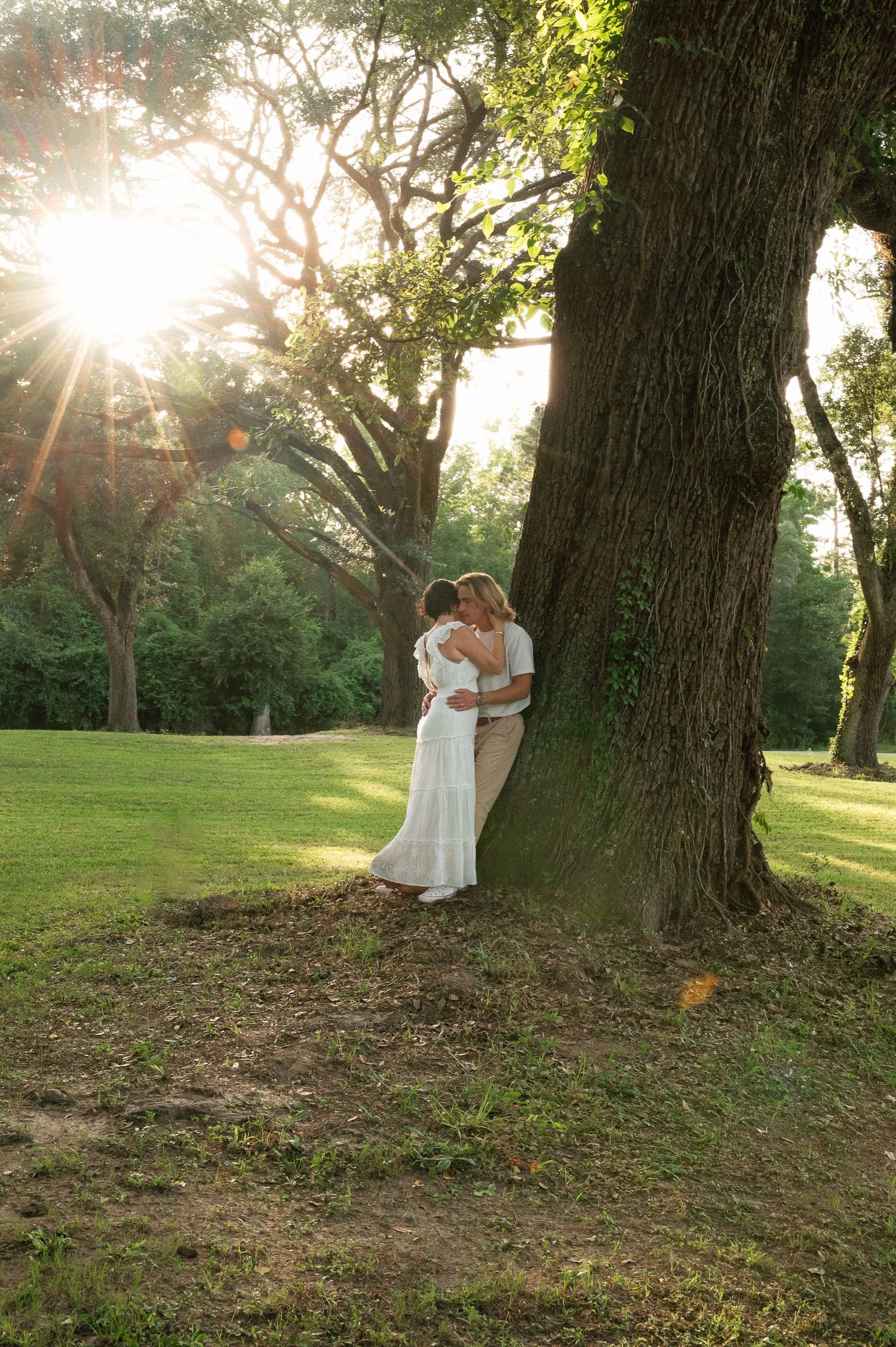 Couple embracing under a large tree at golden hour in a lush green park, bride in white dress.