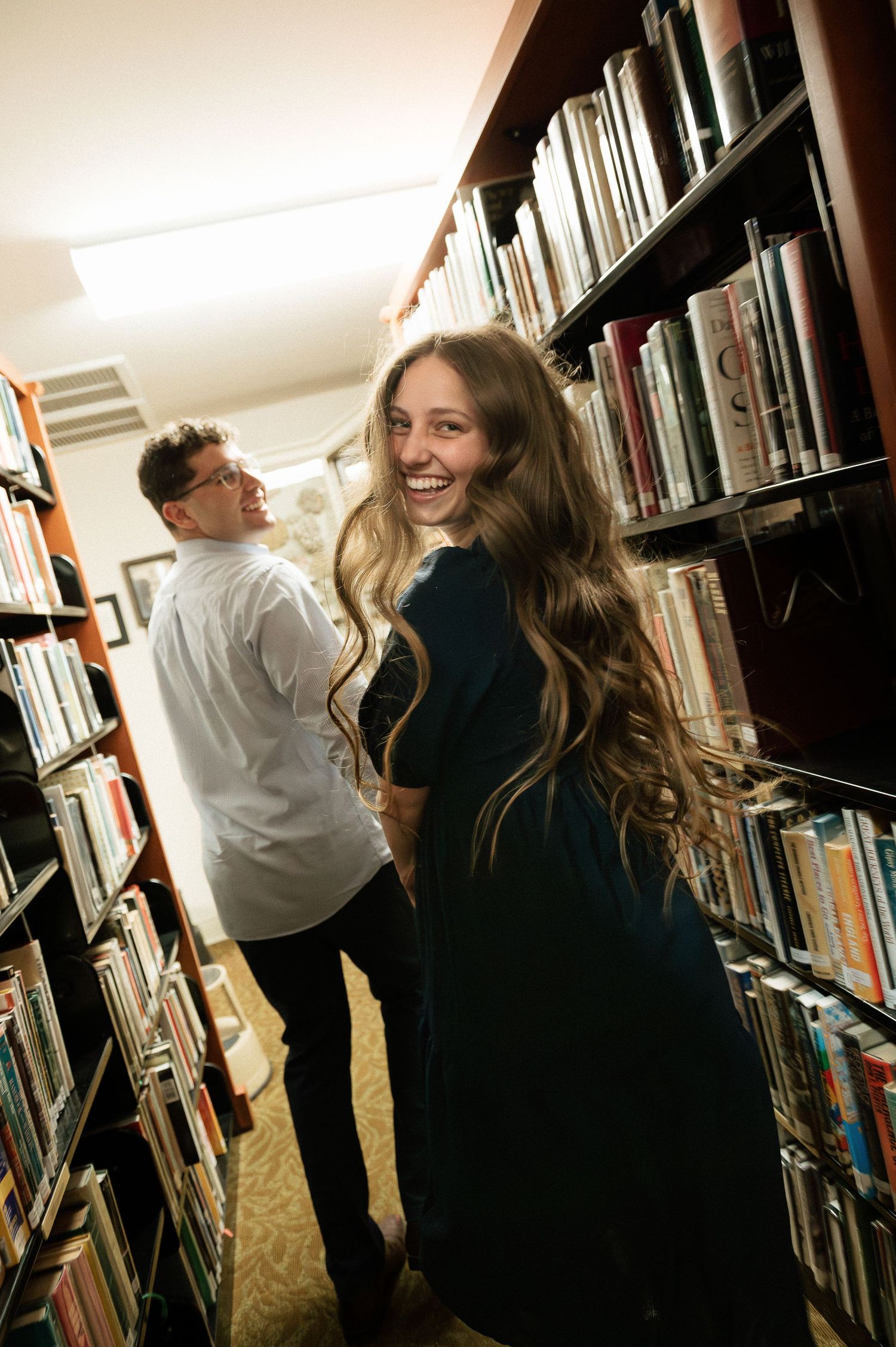 A smiling woman with long wavy hair in a teal dress stands between library bookshelves with a man in white shirt behind her.
