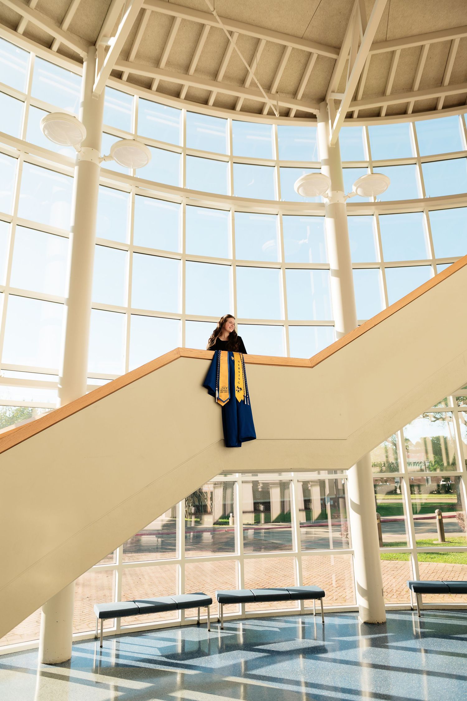 A graduate in blue cap and gown stands atop a grand staircase in a sunlit modern building with floor-to-ceiling windows.