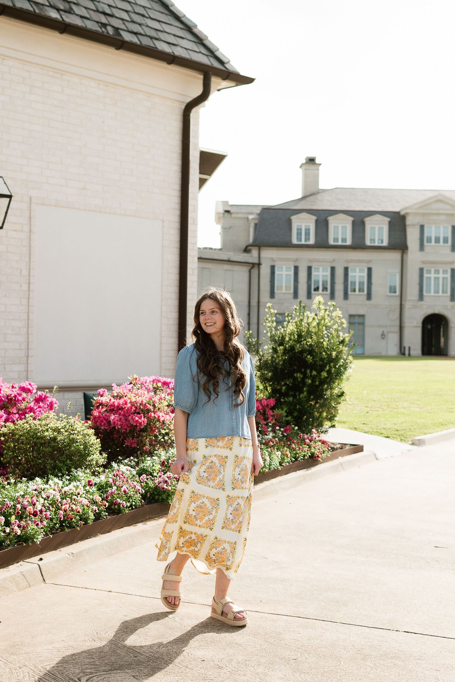 Woman in blue top and floral midi skirt standing on a sunny pathway beside colorful garden blooms near an elegant estate.