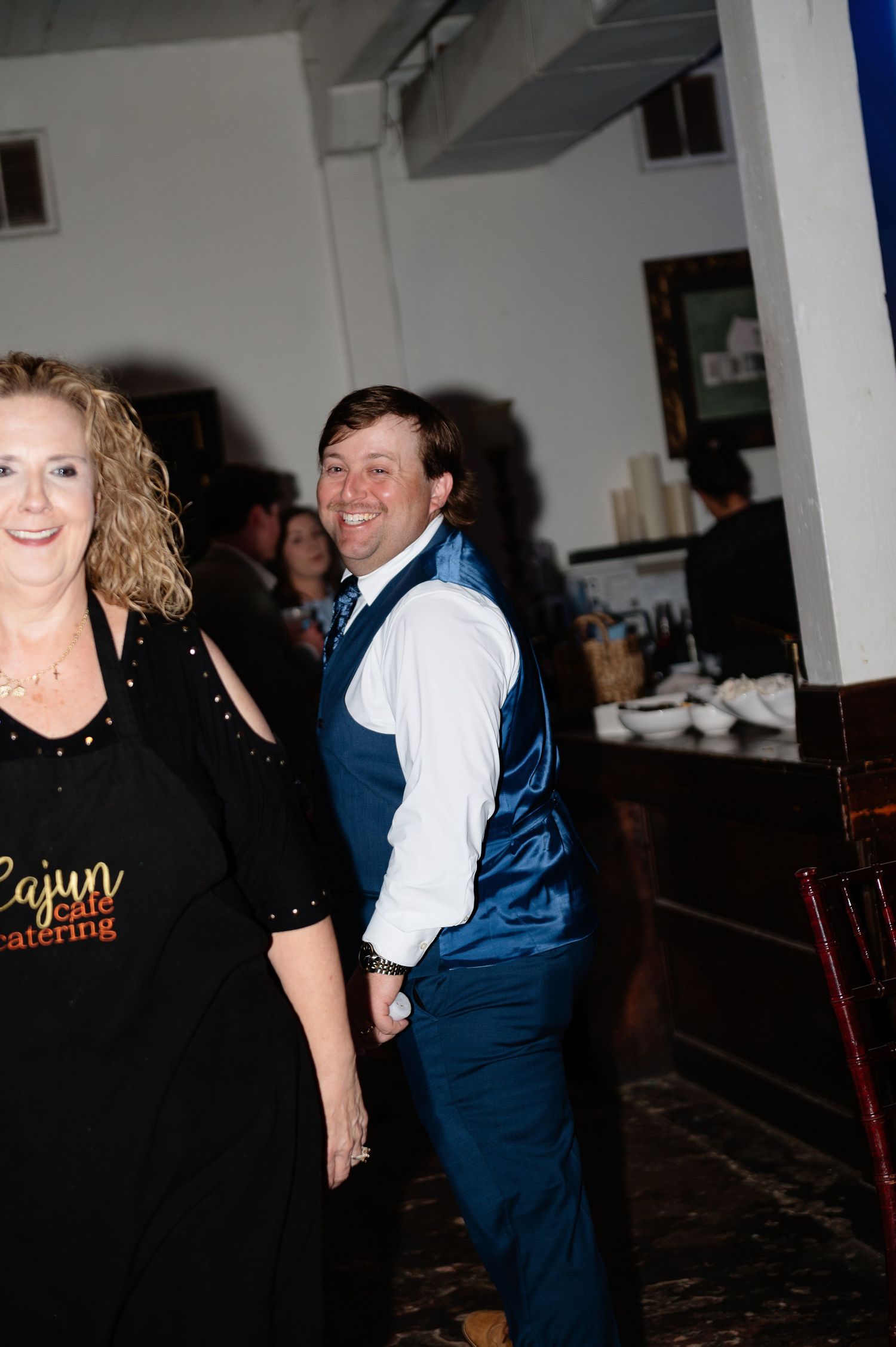 Smiling groom in blue vest and tie at wedding reception with catering staff in background.