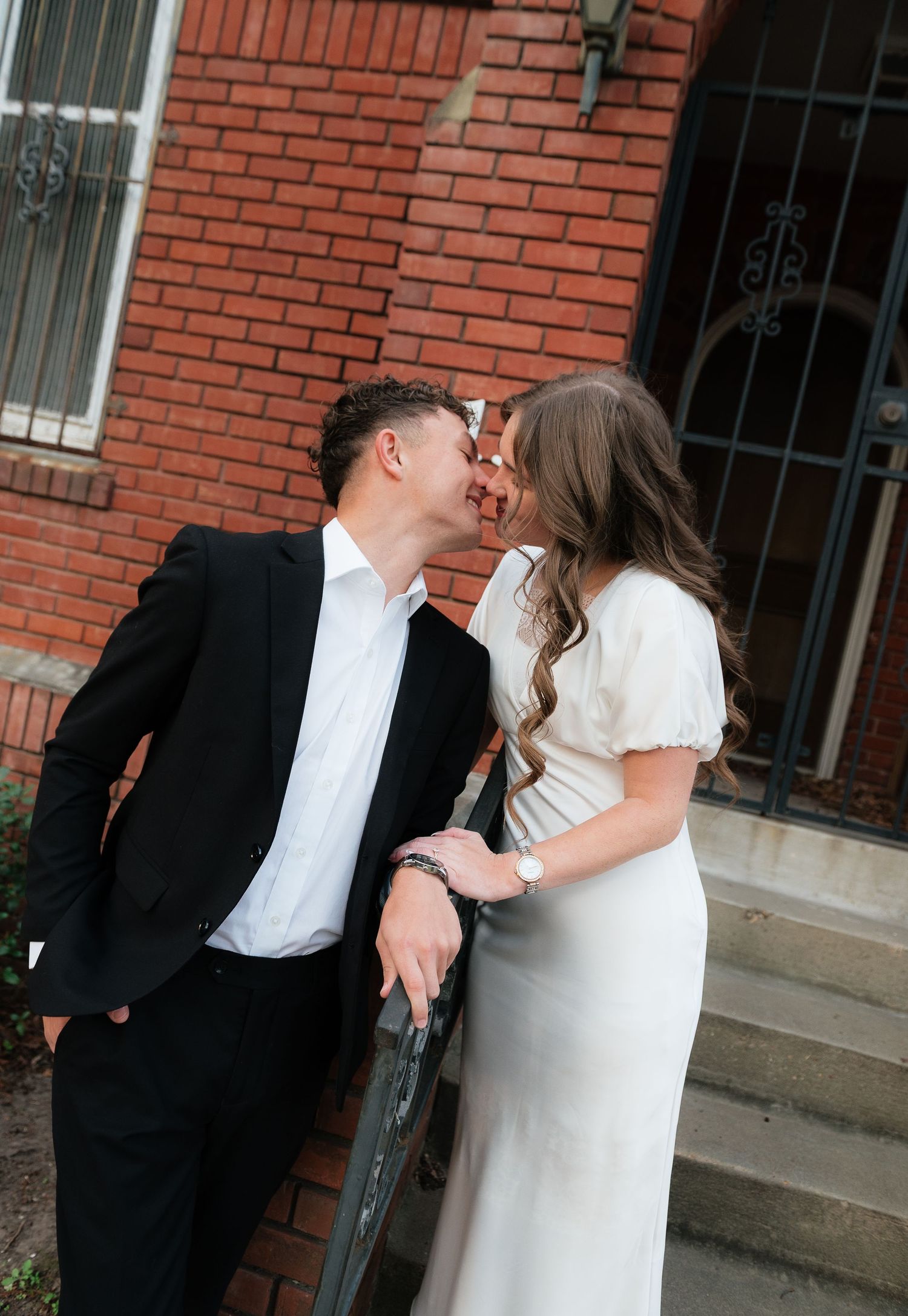 Couple sharing a romantic kiss on brick building steps, man in black suit, woman in white dress.