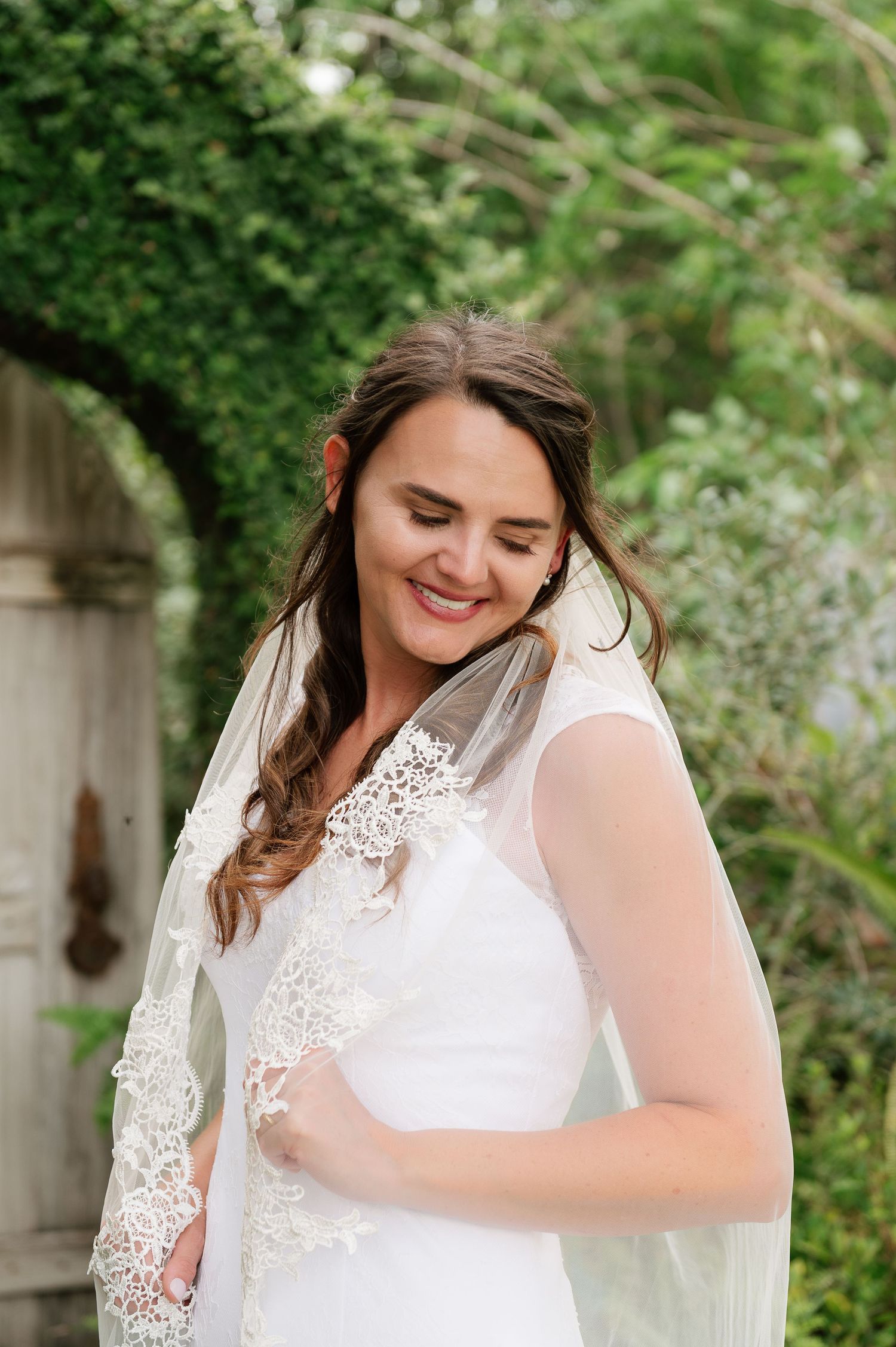 Smiling bride in white lace wedding dress and veil standing in a lush green garden setting.