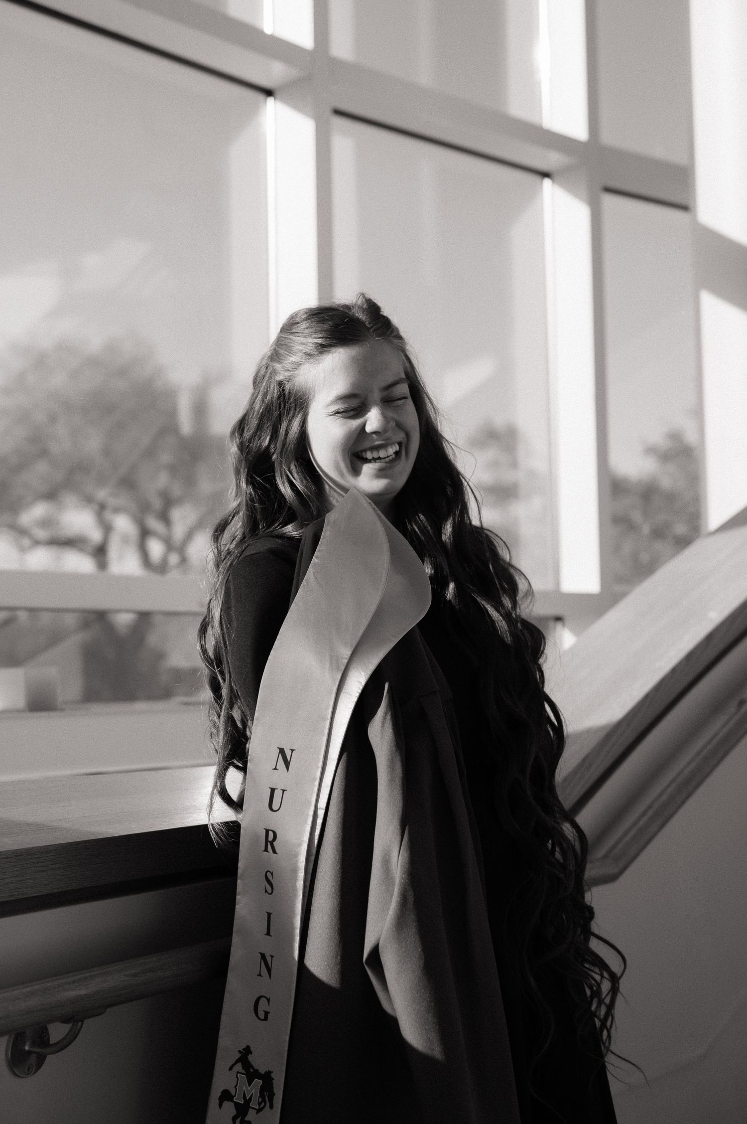 Black and white photo of a smiling nursing graduate wearing a sash and gown near a bright window.