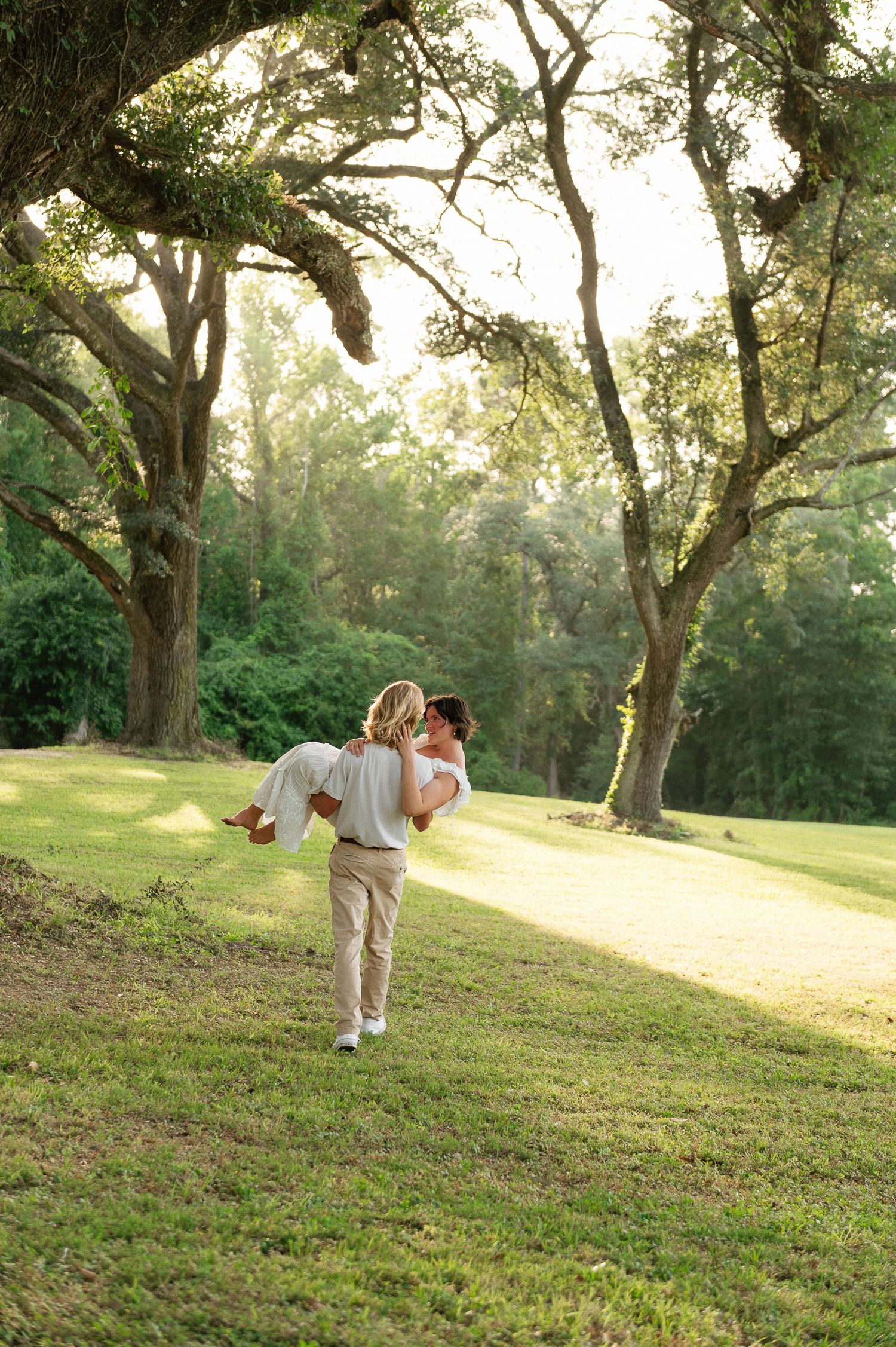 A man lifts his partner in a romantic embrace on a sunlit grassy path surrounded by large oak trees.