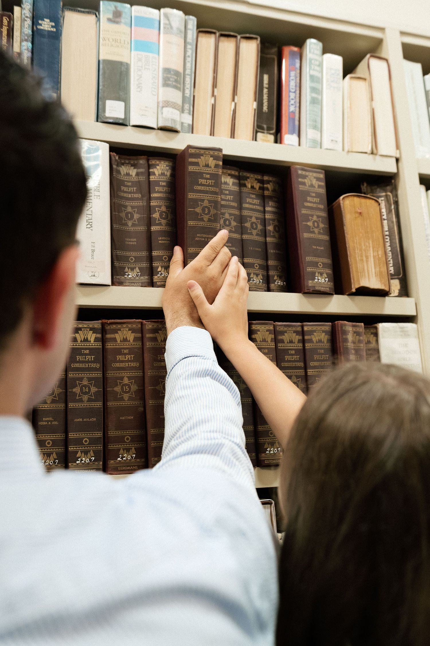 Two people selecting vintage books from a wooden library shelf filled with old leather-bound volumes.