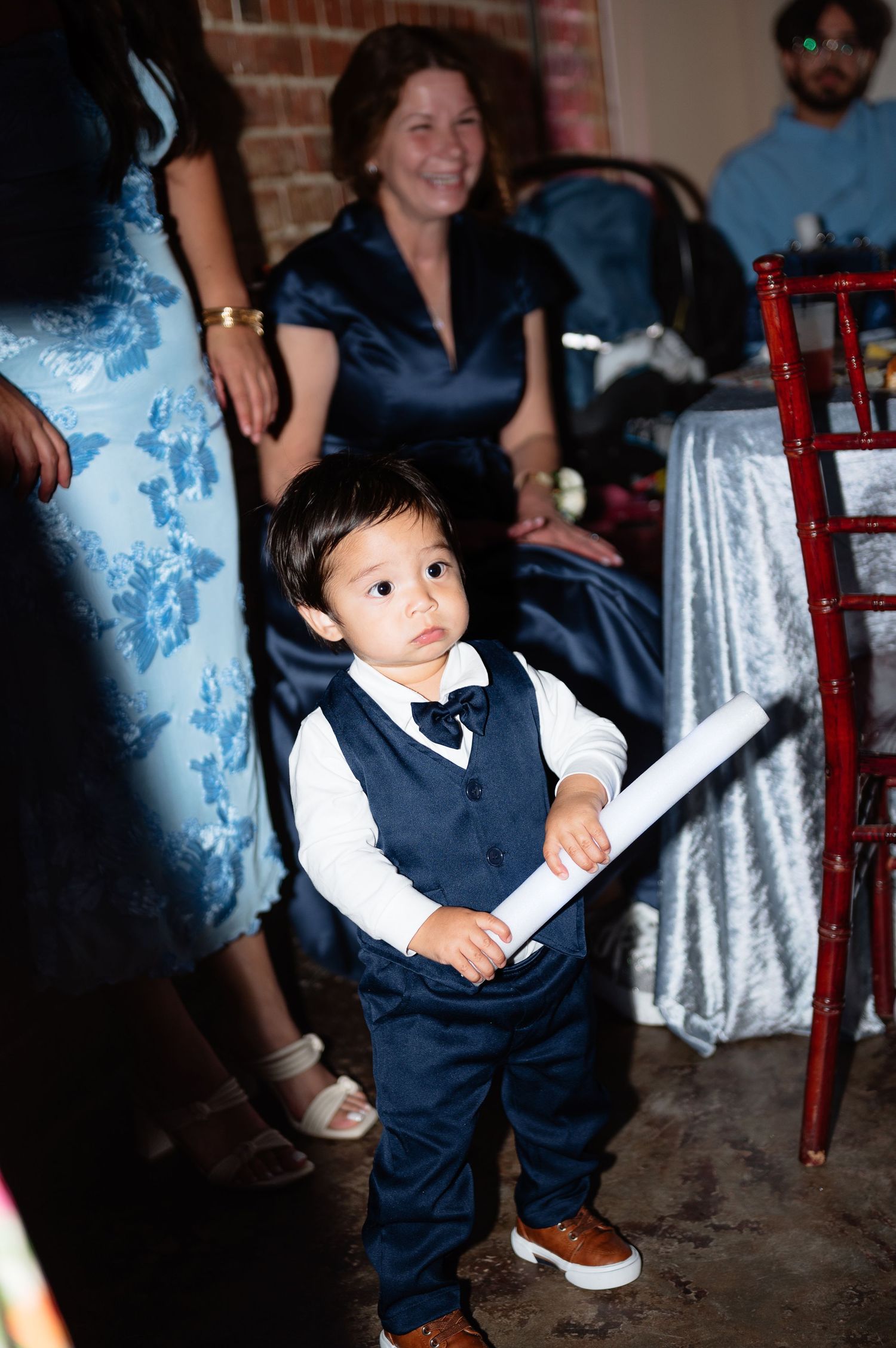 Adorable ring bearer in navy vest and bow tie holding scroll at wedding reception.