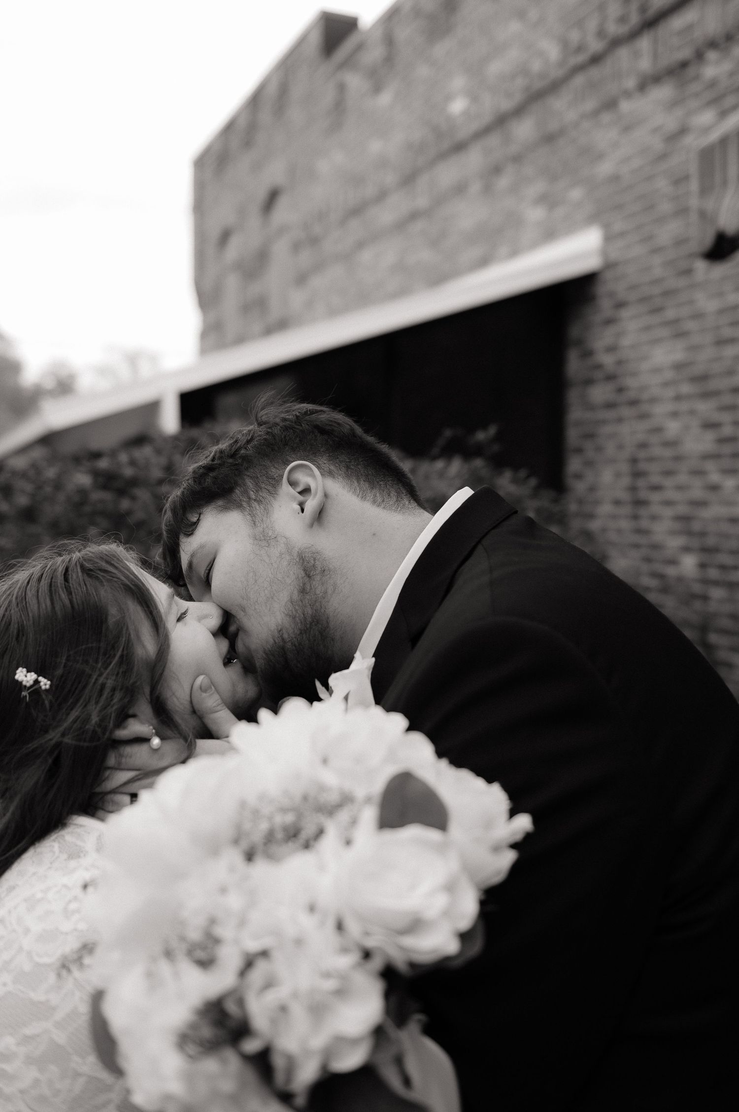 Black and white photo of bride and groom sharing a romantic kiss, bride holding a bouquet of peonies.