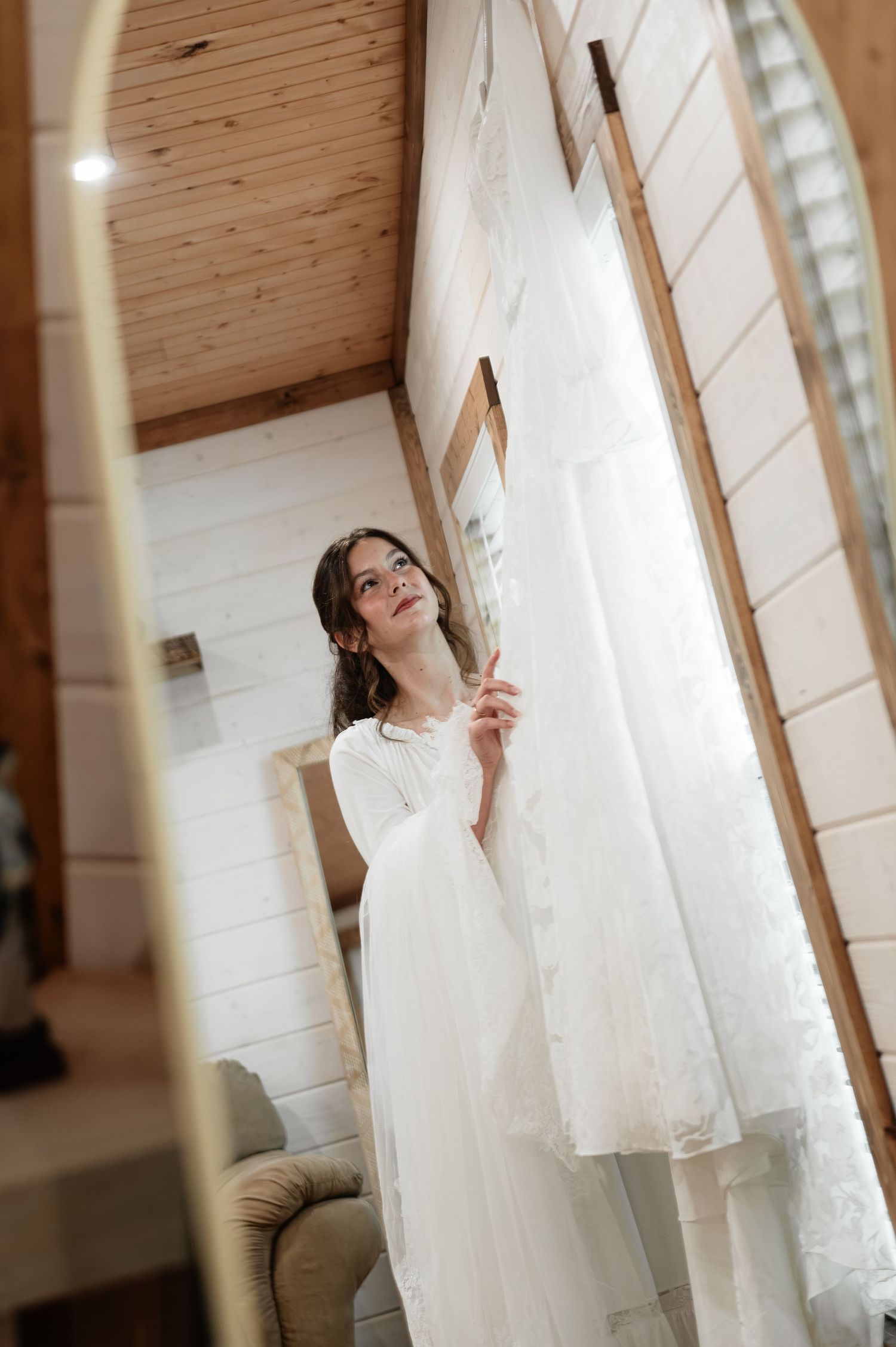 Bride in white gown admiring her wedding dress hanging in a rustic wooden room with natural light.
