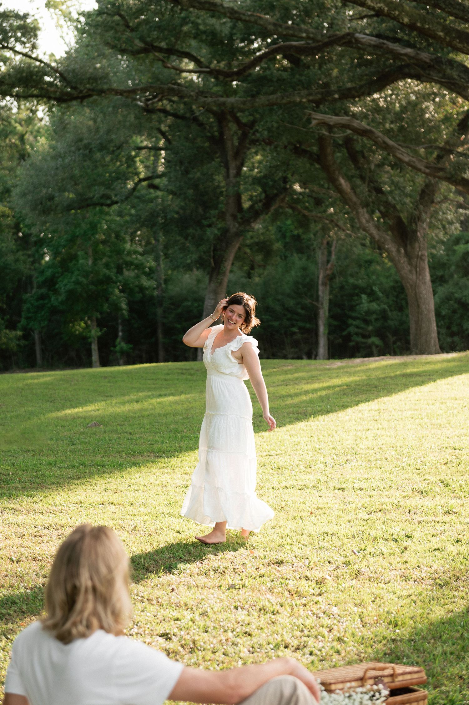 A woman in a white dress walks barefoot on green grass under large oak trees in golden afternoon sunlight.
