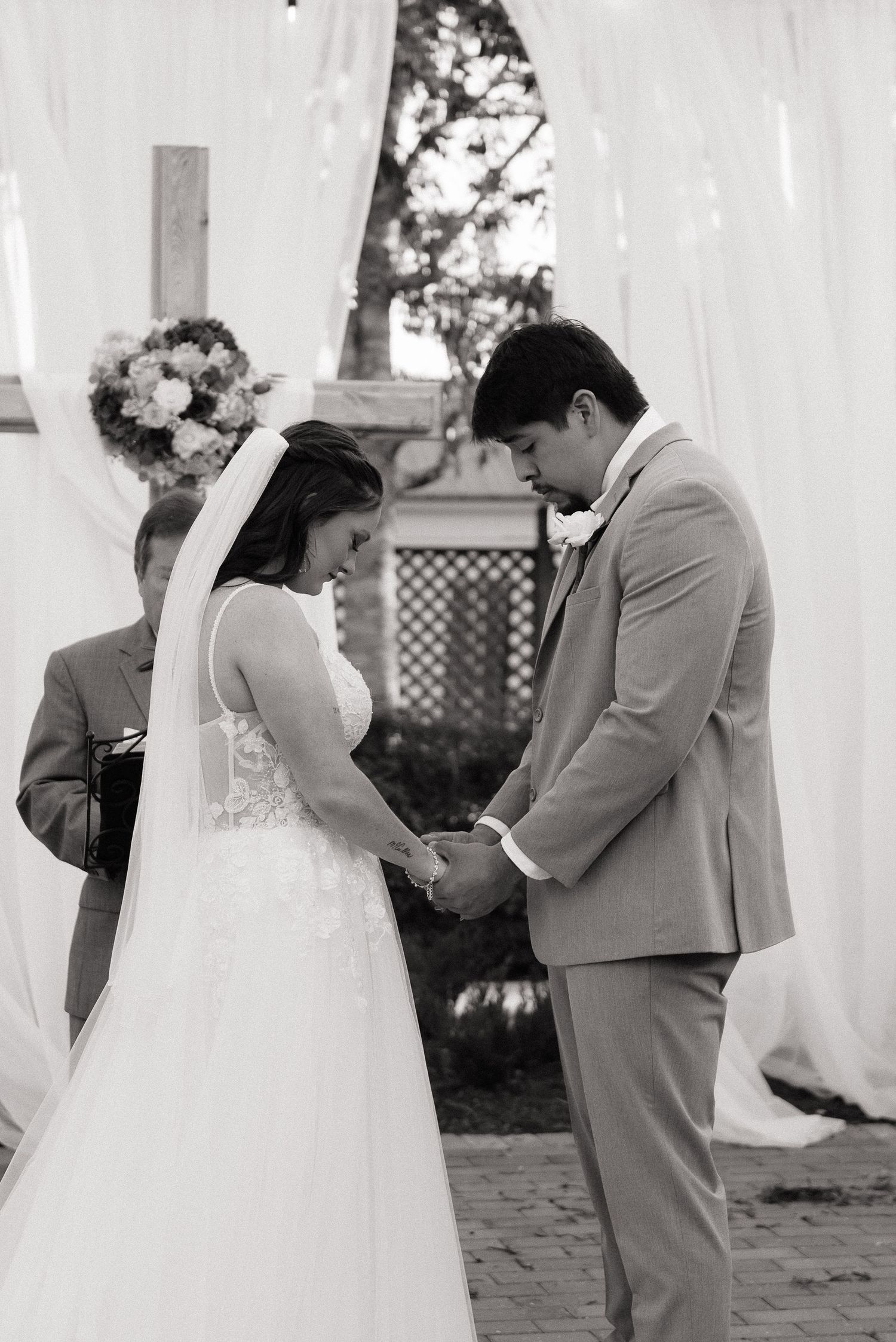 Black and white photo of bride and groom holding hands during outdoor wedding ceremony with floral arch backdrop.