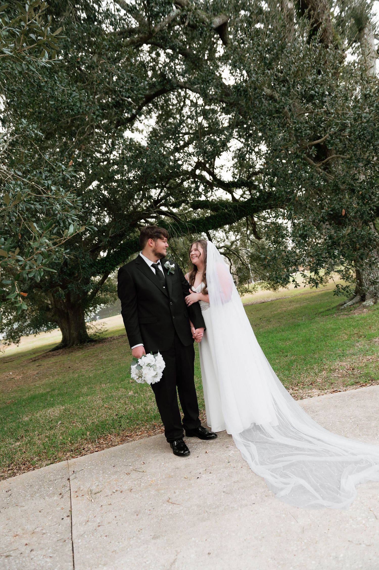 Bride and groom stand beneath a large oak tree on their wedding day, bride wearing a long veil and holding white flowers.