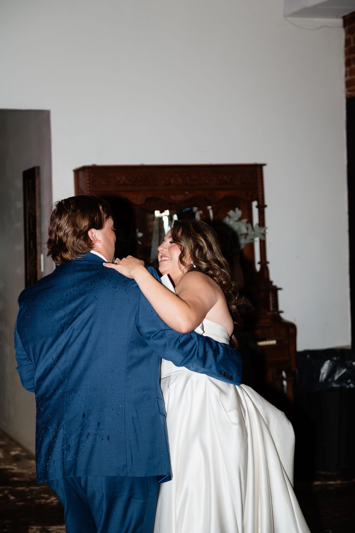 Bride and groom share first dance, bride smiling joyfully in white gown and groom in navy suit.