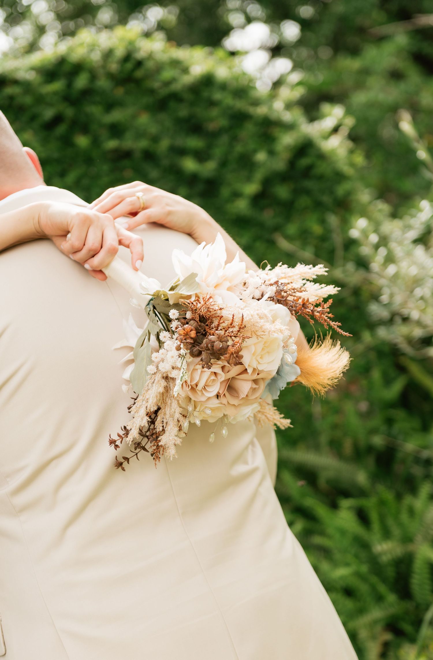 Bride holding a boho dried floral bouquet with roses and pampas grass while embracing groom outdoors.