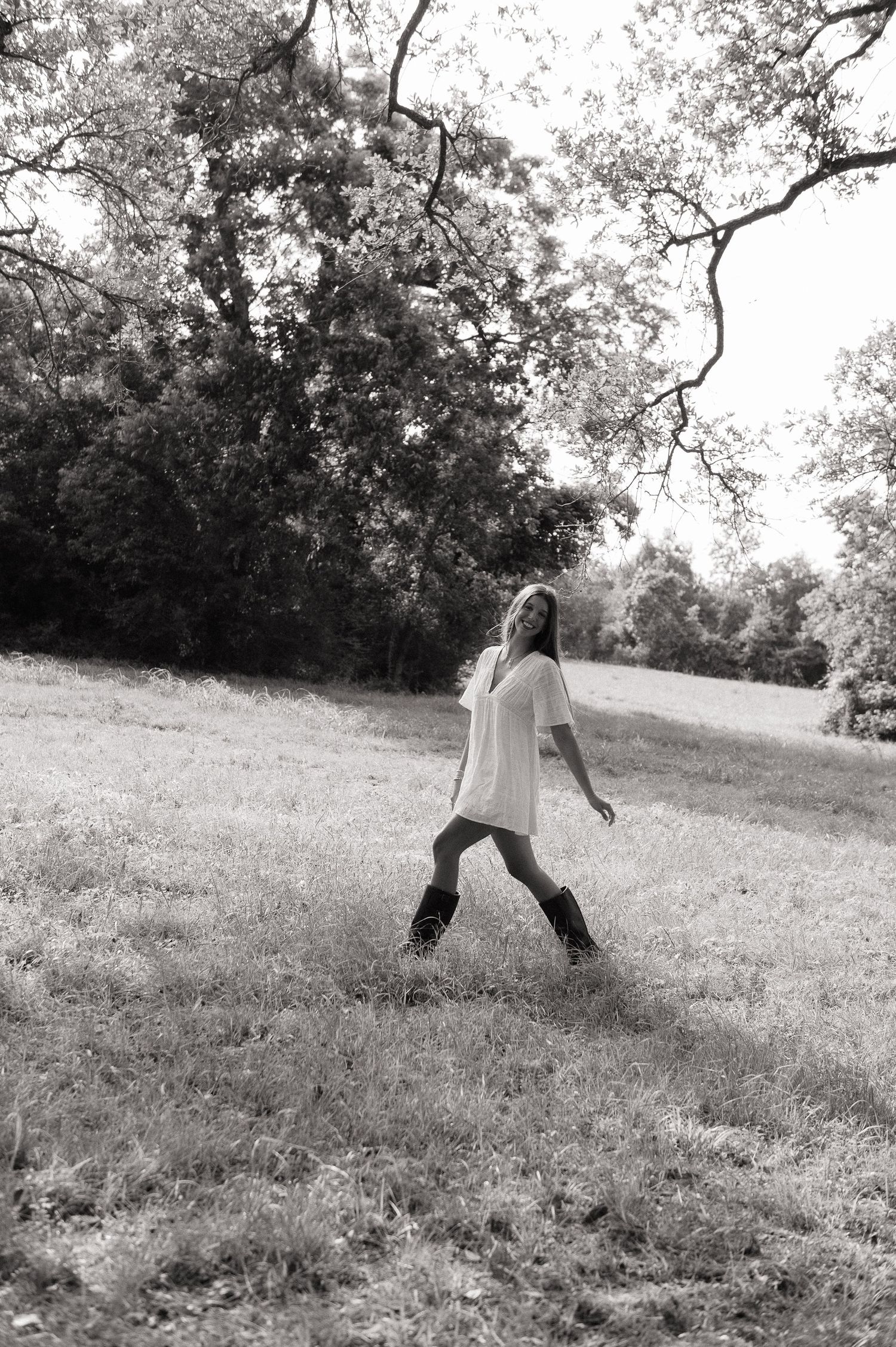 Black and white photo of a young woman in a white tunic and boots walking through a grassy field with trees.
