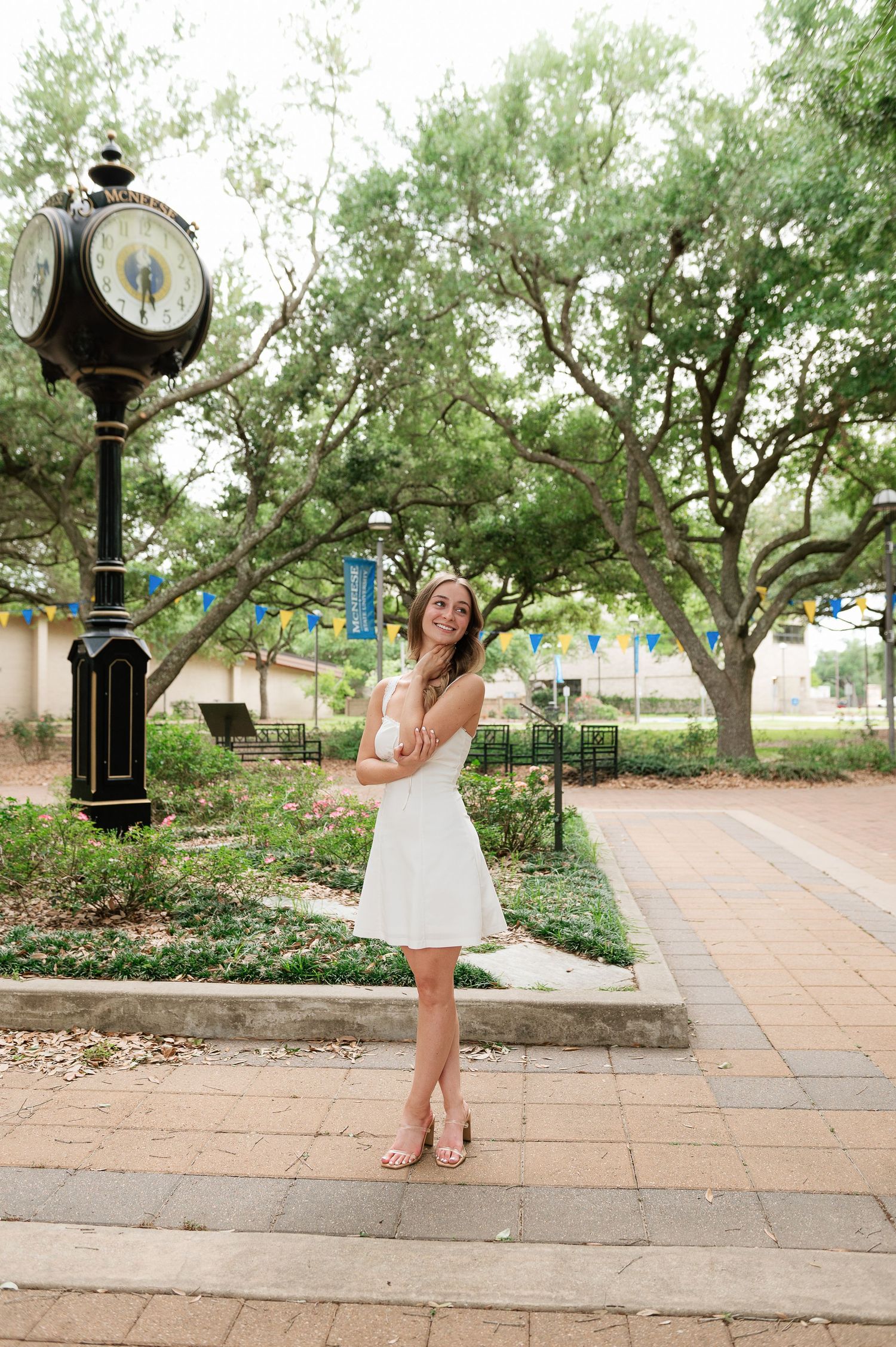 Young woman in white dress posing by vintage street clock on brick walkway surrounded by green trees.