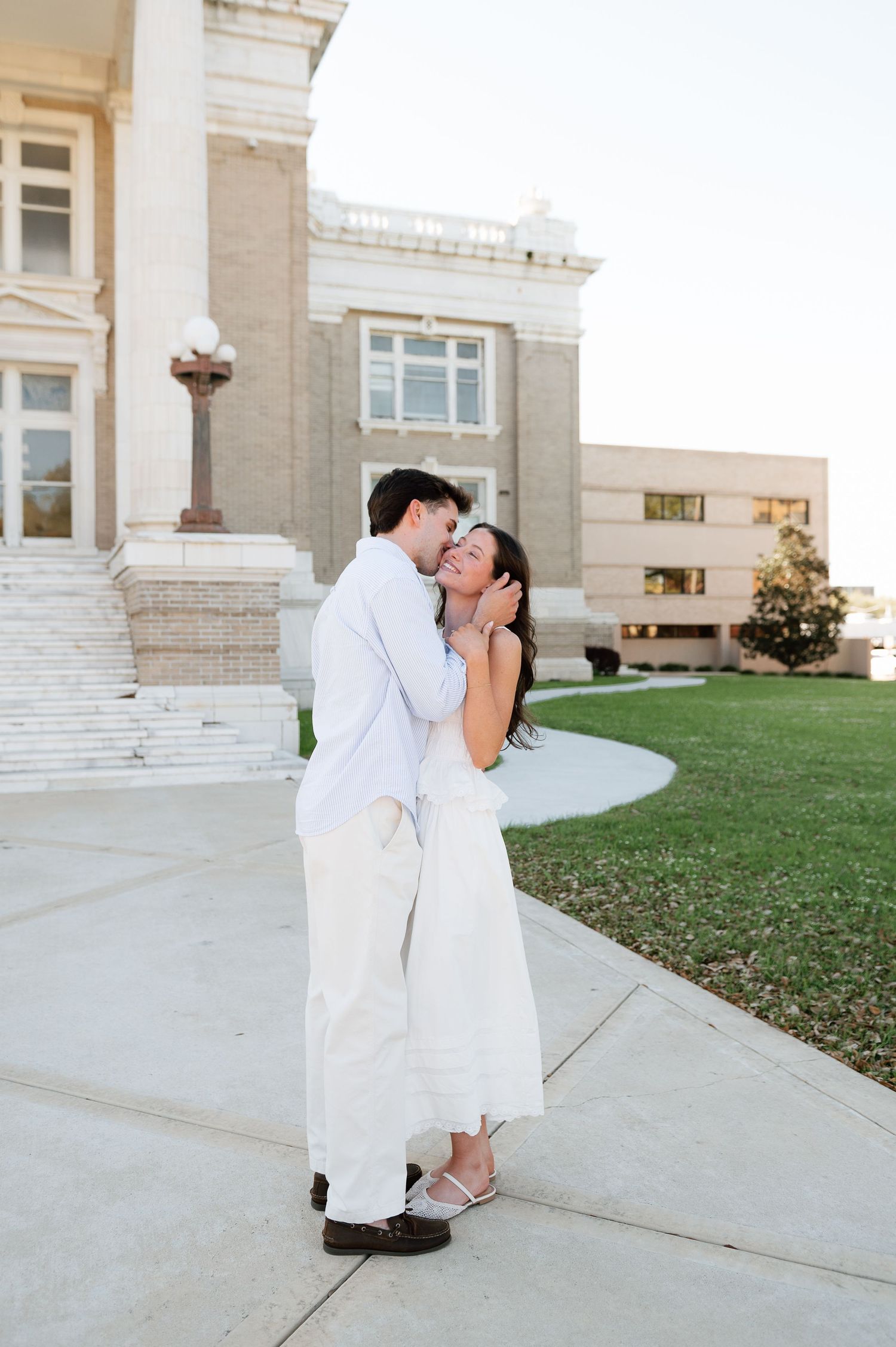 Couple embracing and kissing in front of a classical building, both dressed in white outfits on a sunny day.